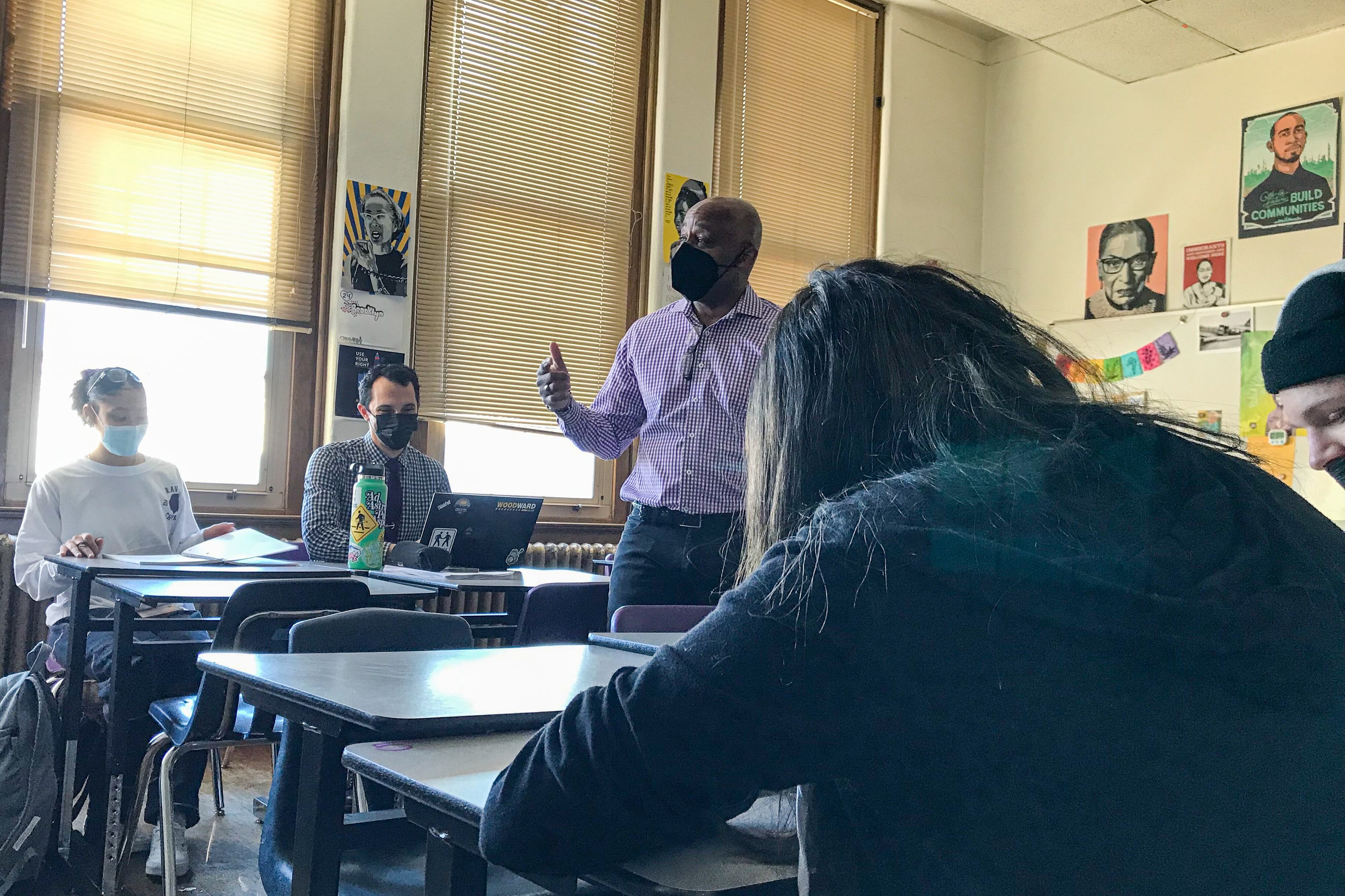 Denver Mayor Michael Hancock, wearing a purple and white shirt, teaches in a high school classroom while students listen. He’s wearing a face mask and gesturing.