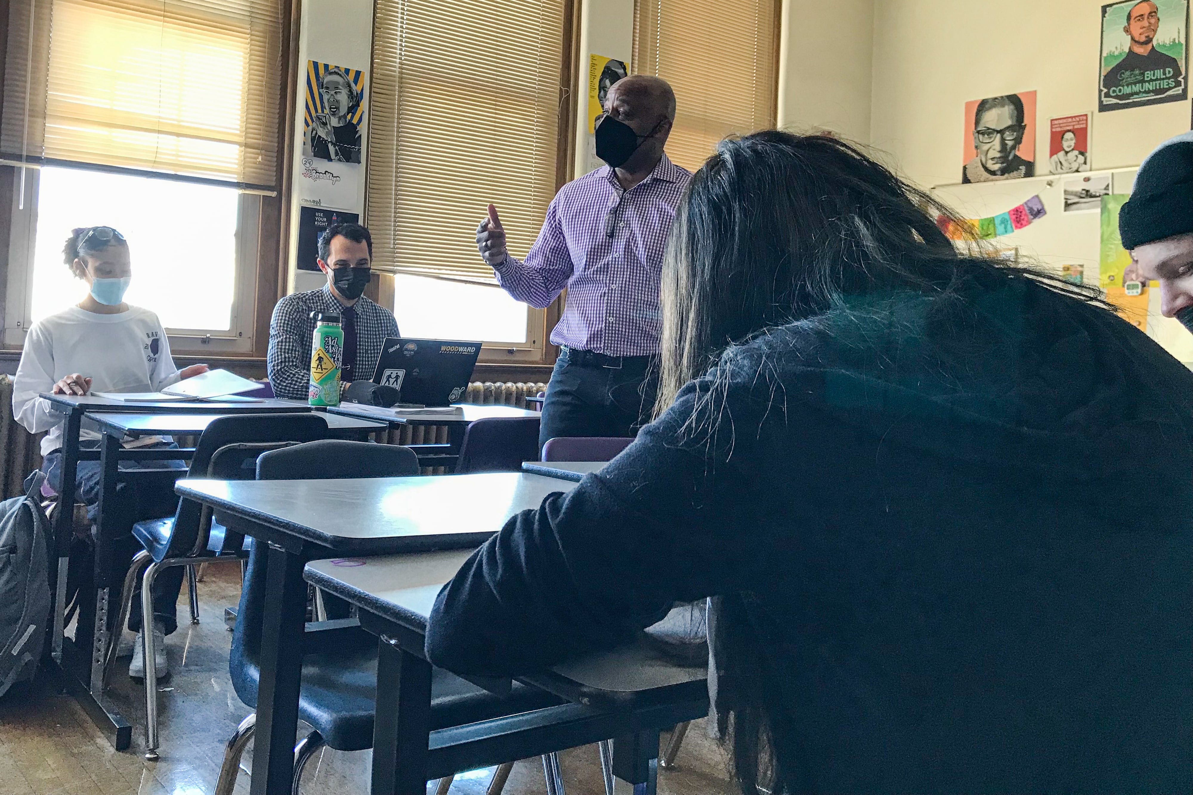 Denver Mayor Michael Hancock, wearing a purple and white shirt, teaches in a high school classroom while students listen. He’s wearing a face mask and gesturing.