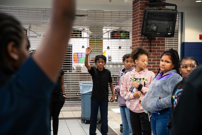 A middle school boy wearing all black raises his hand while four students stand to his left. There is a wire gate that blocks off the kitchen while they stand in a school cafeteria.