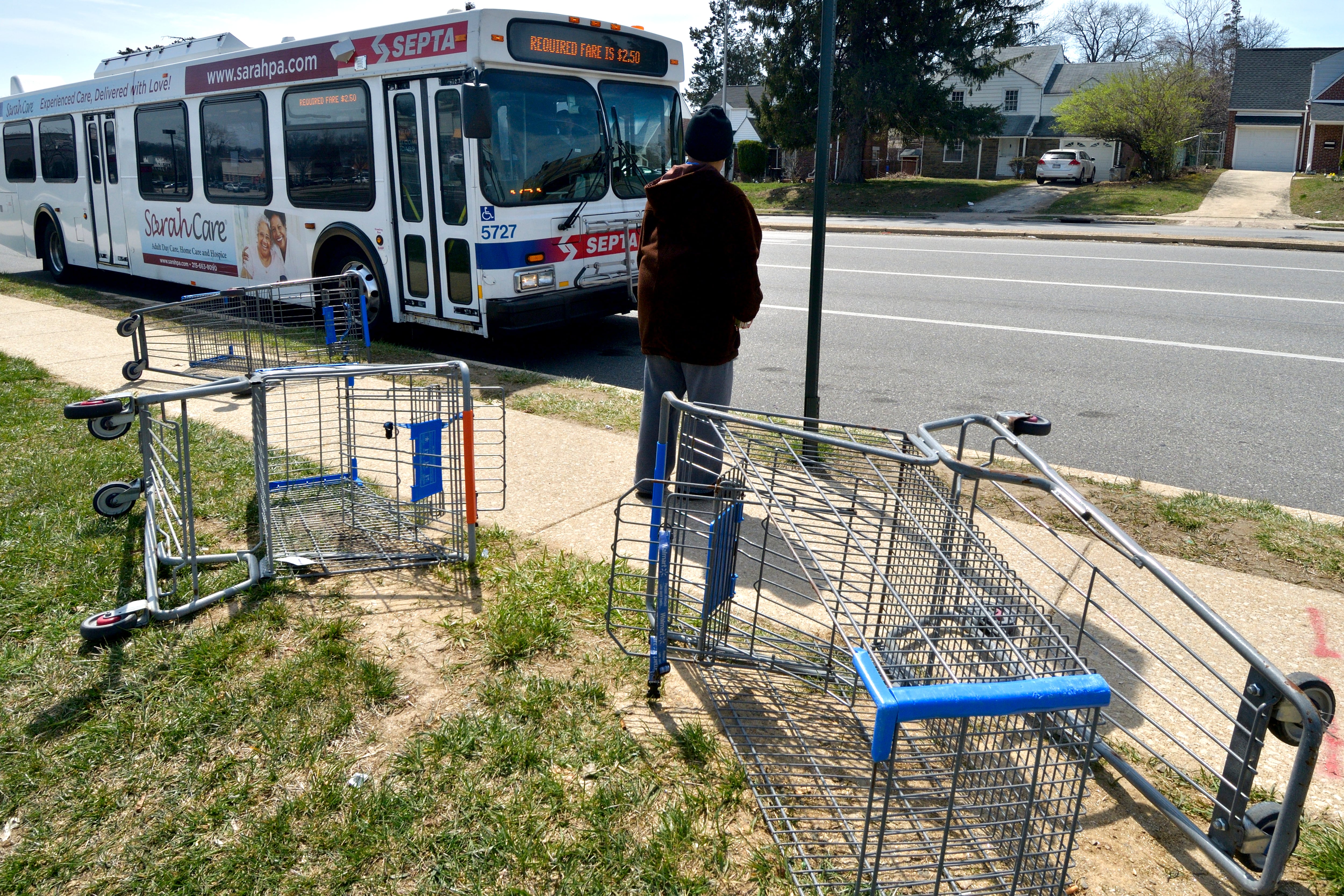 A pedestrian waits on the side of the road for a SEPTA bus. Three abandoned shopping carts sit in the grass near the sidewalk at the bus stop.