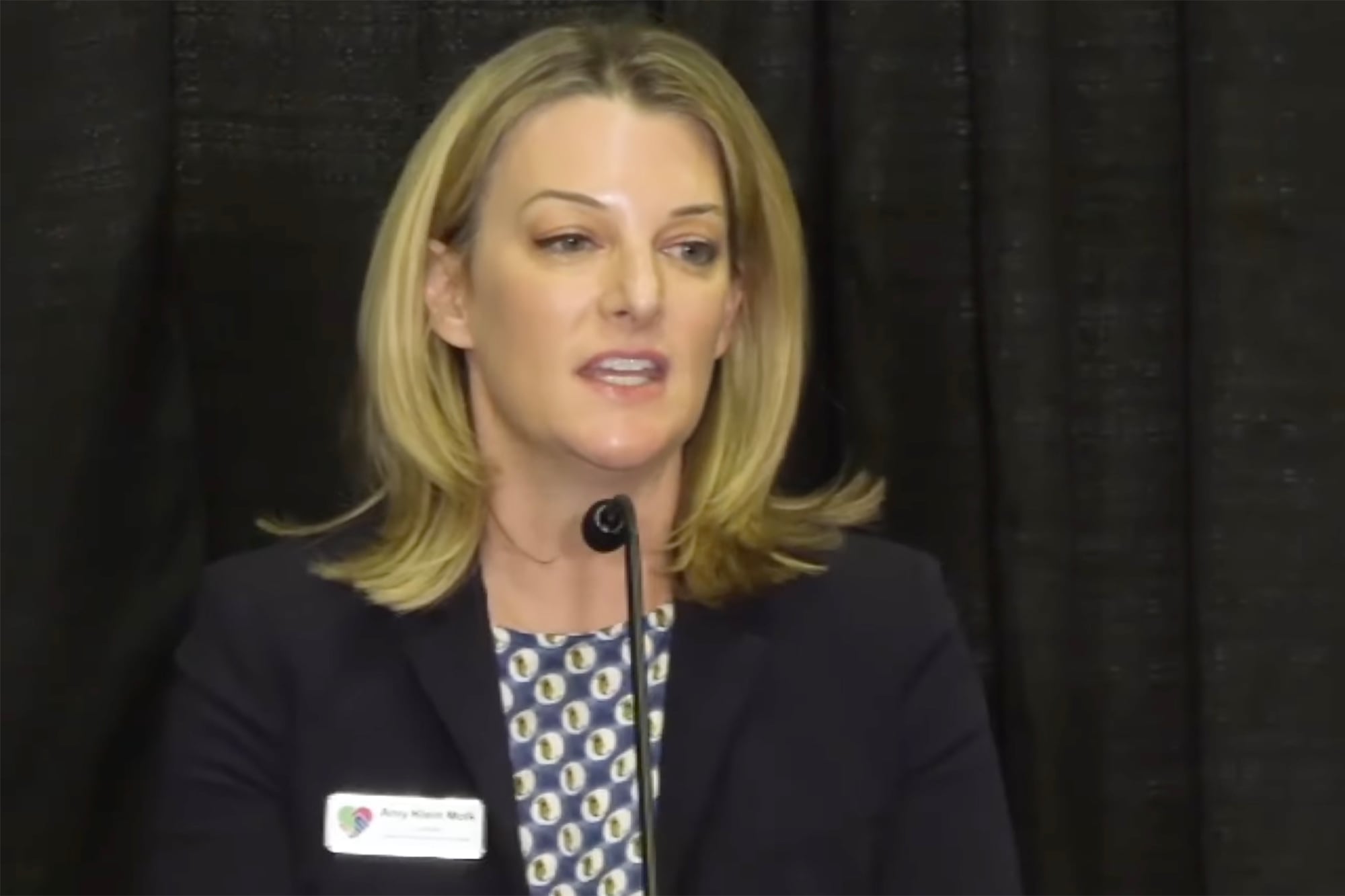 A screen grab of a white woman with blonde hair wearing business clothes and a small white name tag speaking into a small microphone with a dark background.