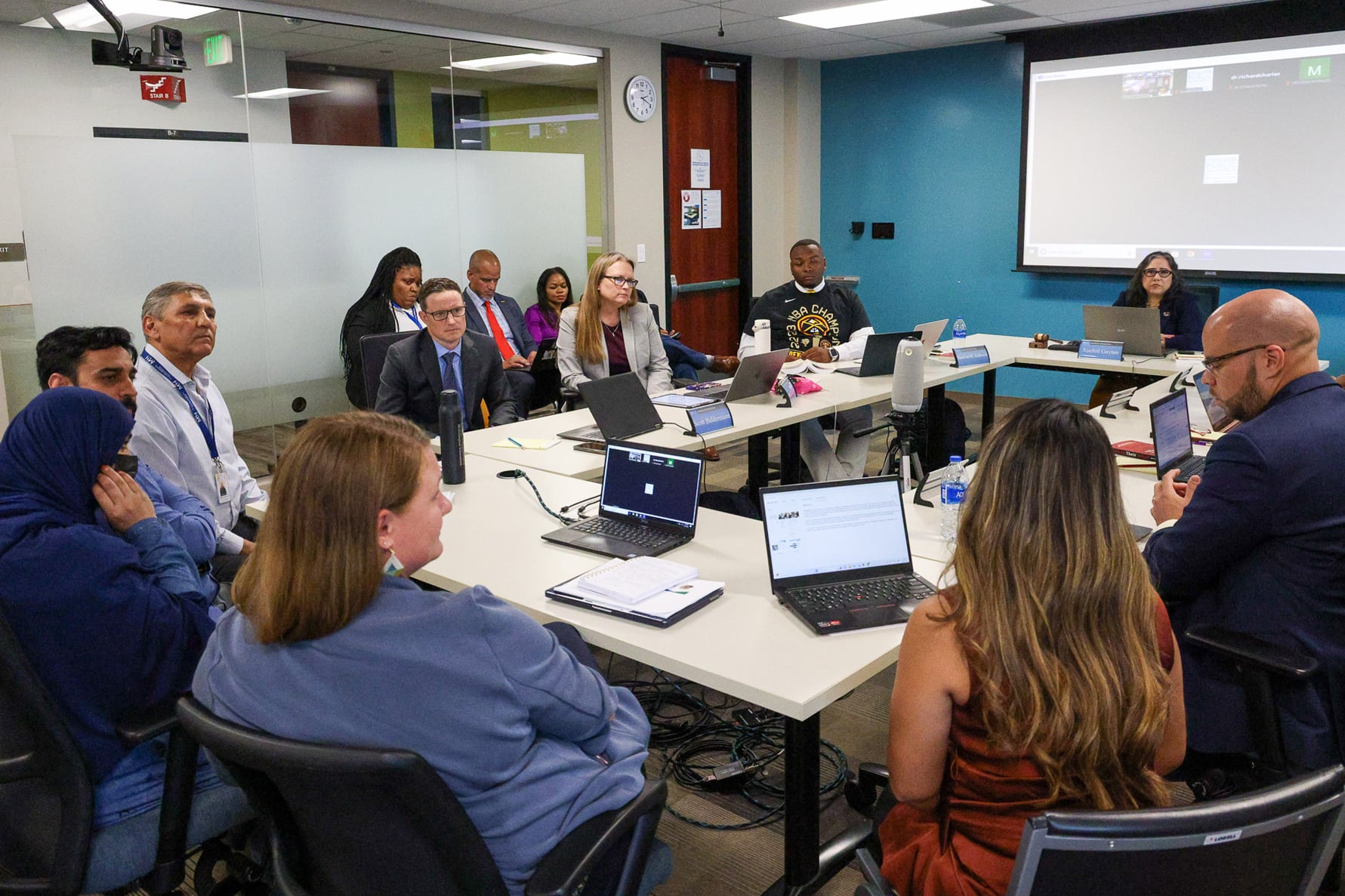 A large group of people sit at tables that are in the shape of a rectangle in a room.