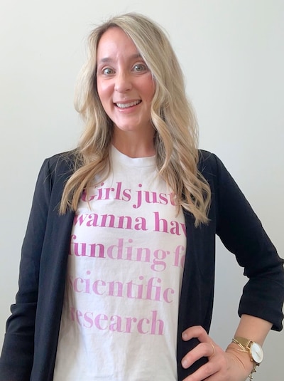 A woman with blond hair wears a t-shirt that says, "Girls just wanna have funding for scientific research." She is wearing a black blazer.