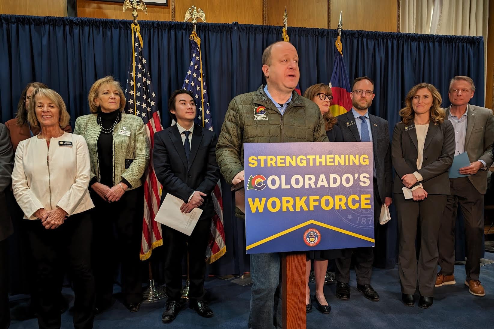 A man wearing a green jacket stands at a podium with a sign that reads "Strengthening Colorado's workforce." There is a group of people standing in a row with flags in the background.
