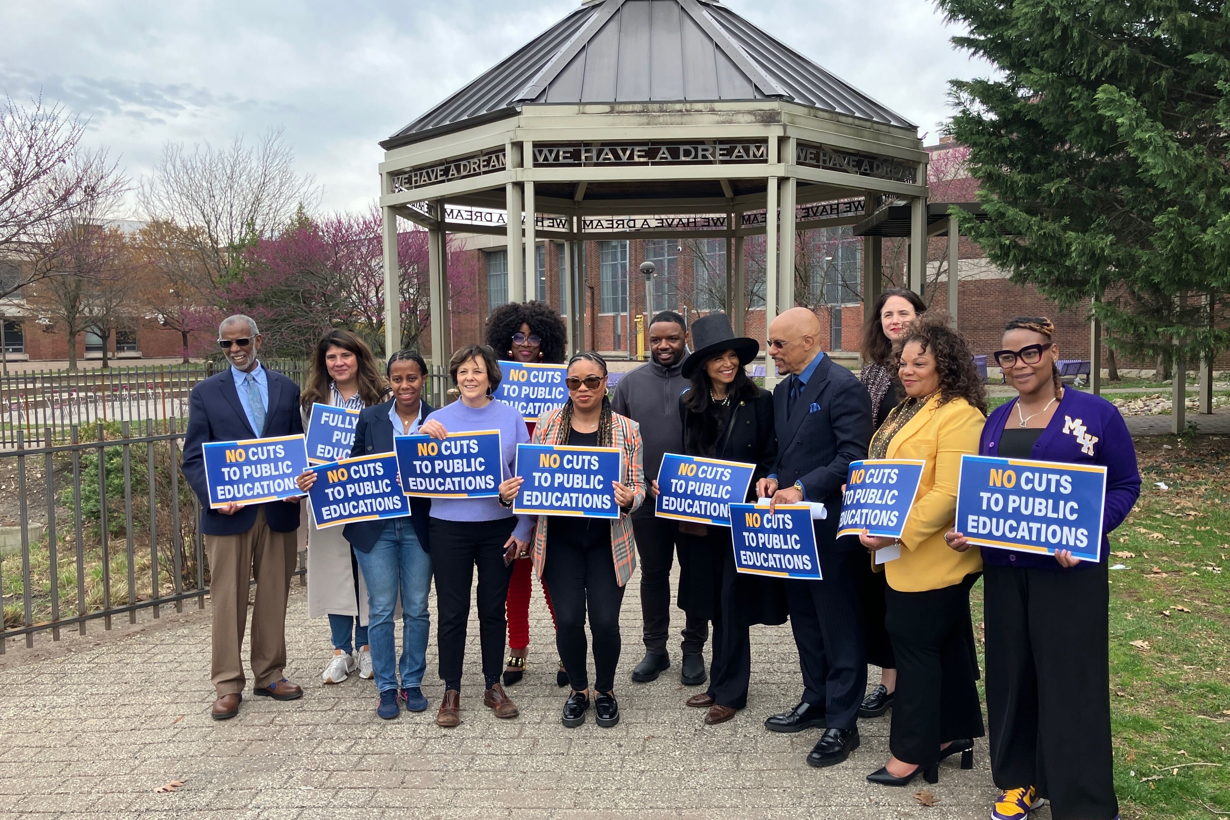 A group of officials and advocates stand in a courtyard and hold signs that read "no cuts to public educations."