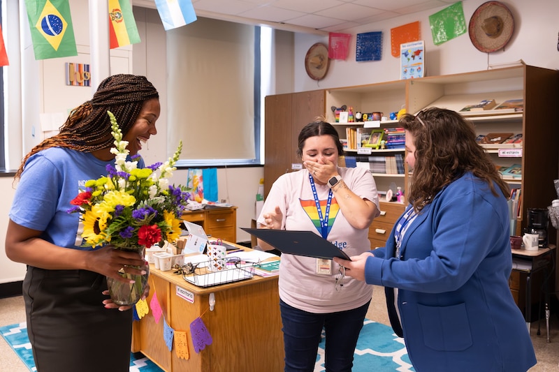 Three adults stand next to each other while the two adults on each end surprise the middle adult with an award and flowers.