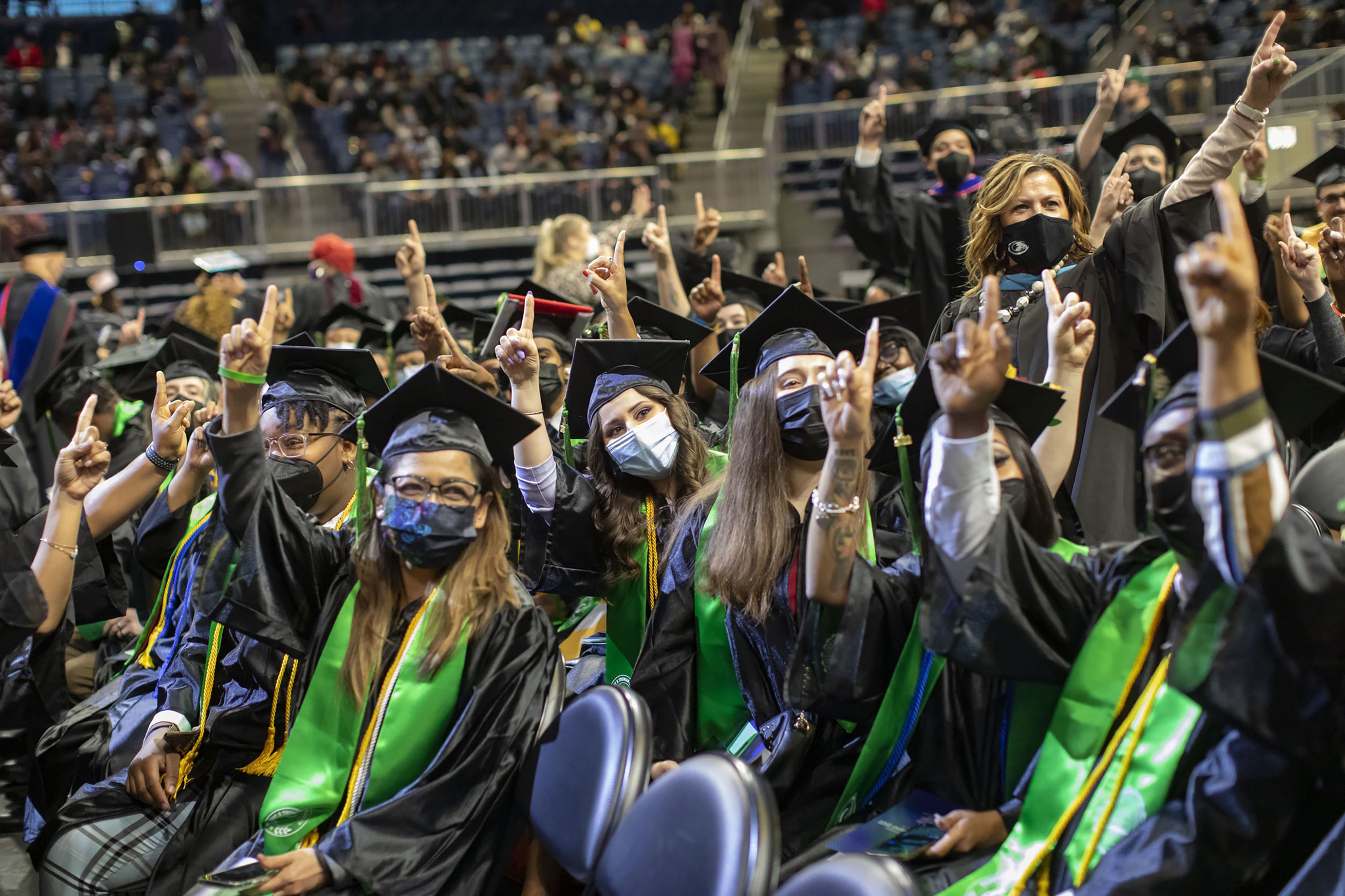 Young people wearing graduation caps and gowns point fingers up in the air.