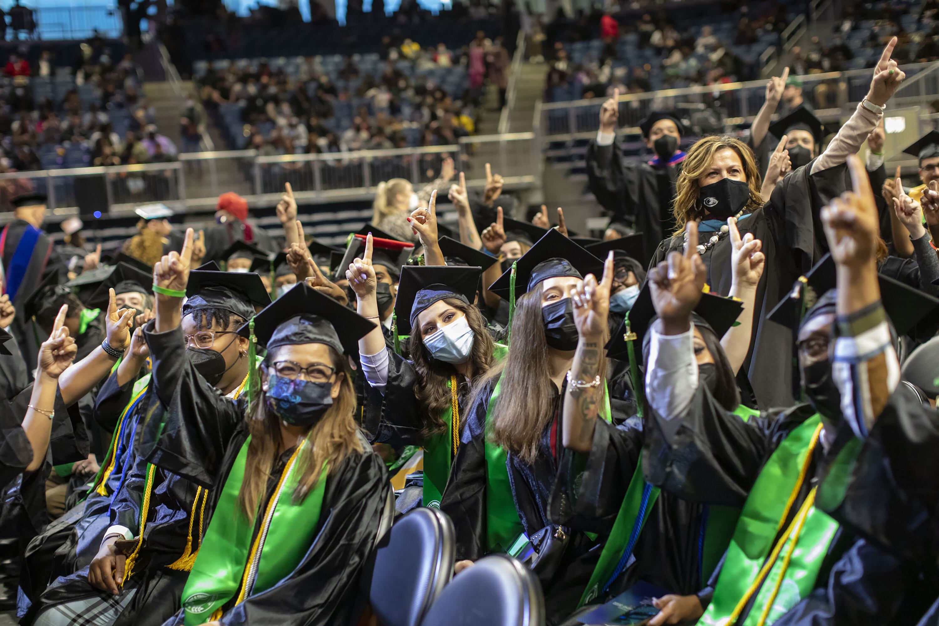 Young people wearing graduation caps and gowns point fingers up in the air.