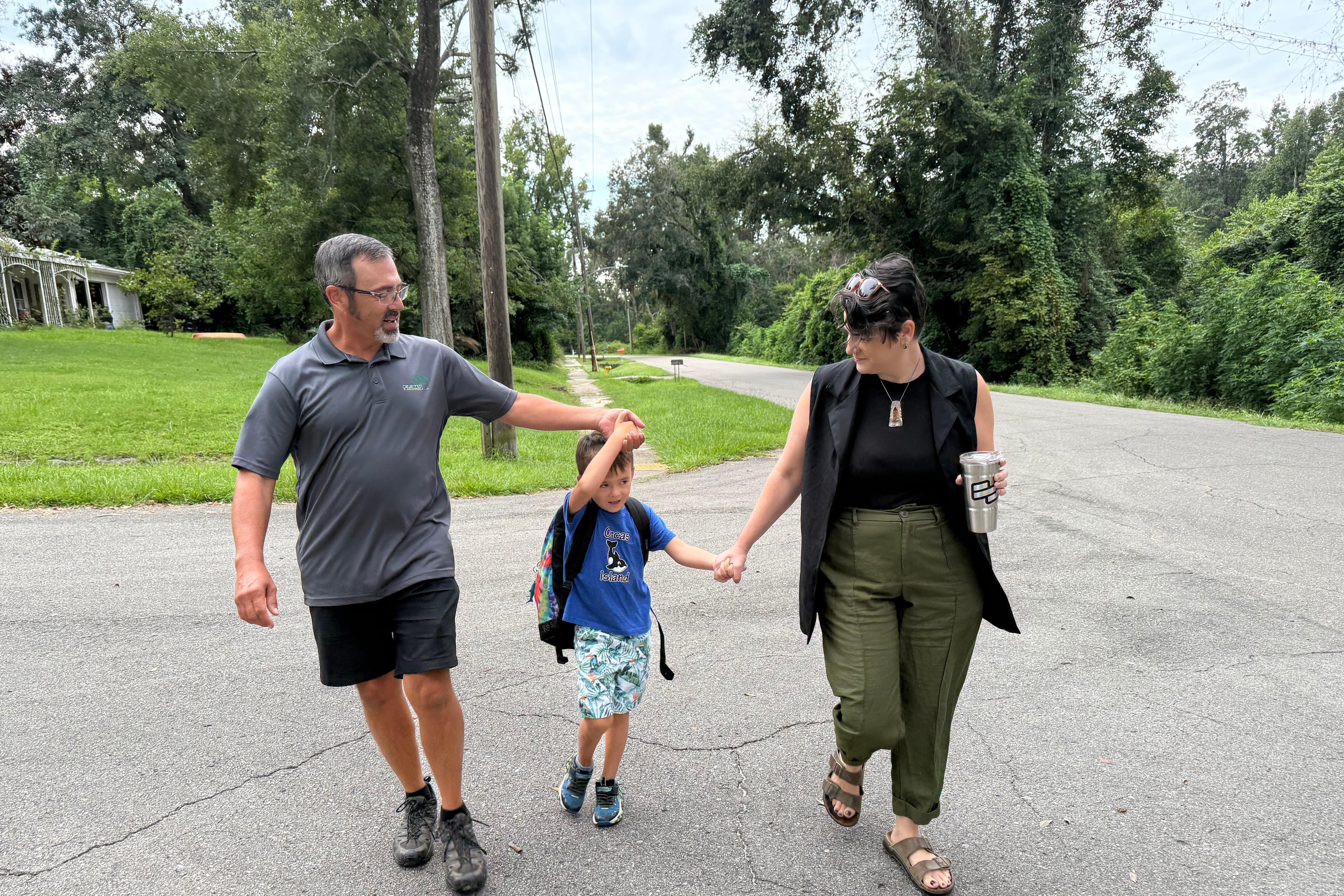 A photograph of two white parents each holding the hand of a small child carrying a backpack outside.