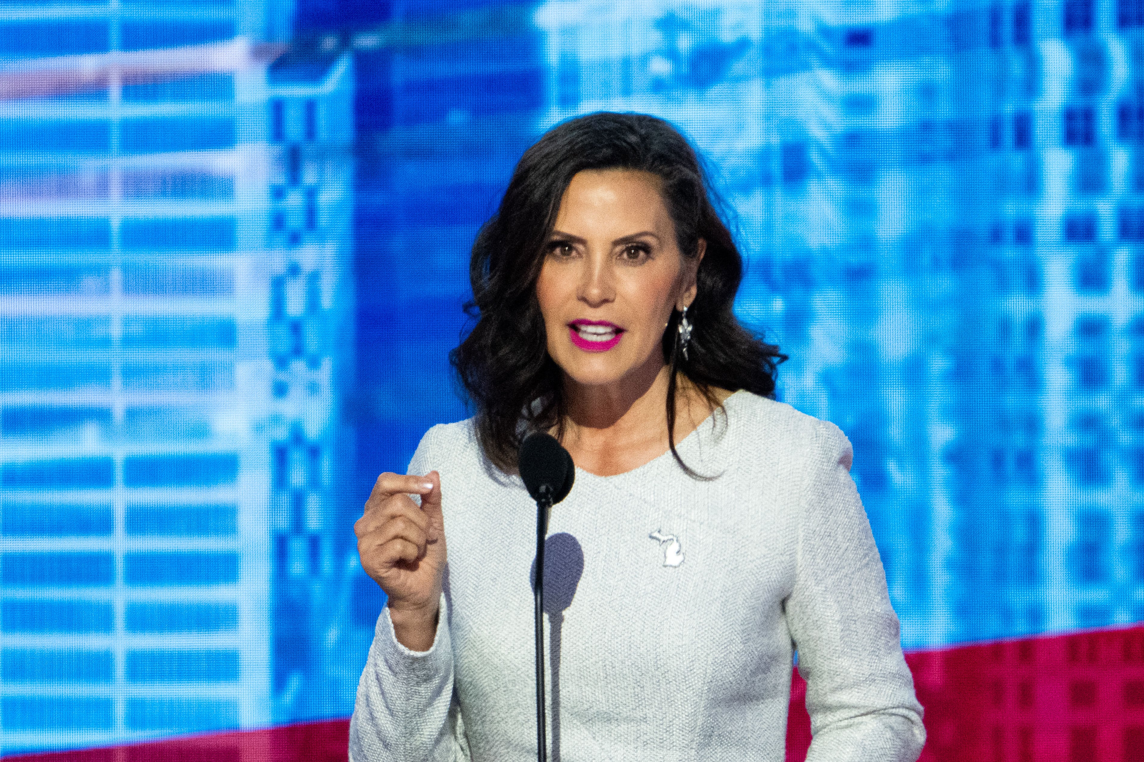 A woman with short dark hair and wearing a light colored blouse speaks from behind a small microphone in front of a blue and red background.