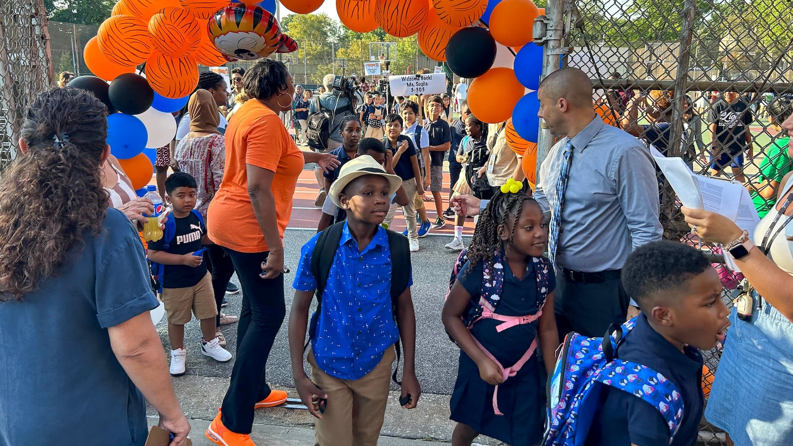 A child in a blue shirt walks under an archway of blue and orange balloons, with other people in the background.