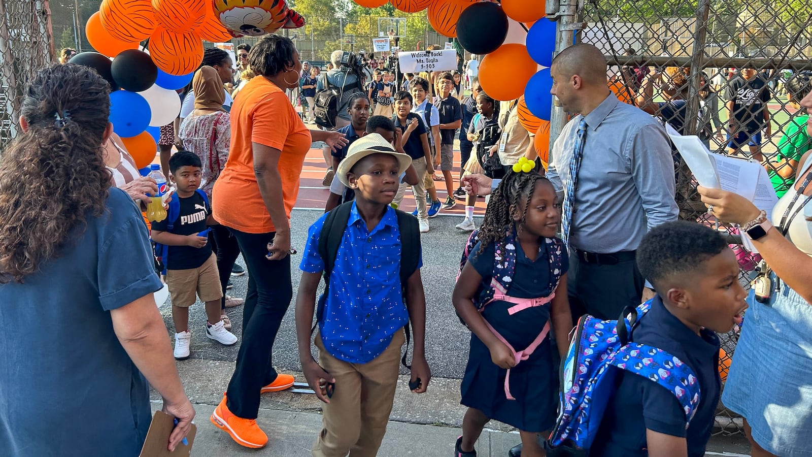A child in a blue shirt walks under an archway of blue and orange balloons, with other people in the background.