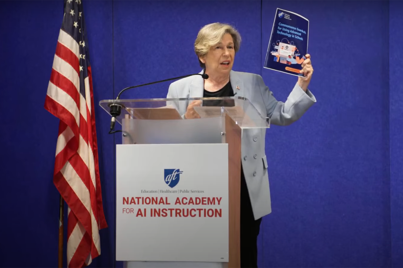 A woman in a light jacket stands next to a flag and holds up a paper.