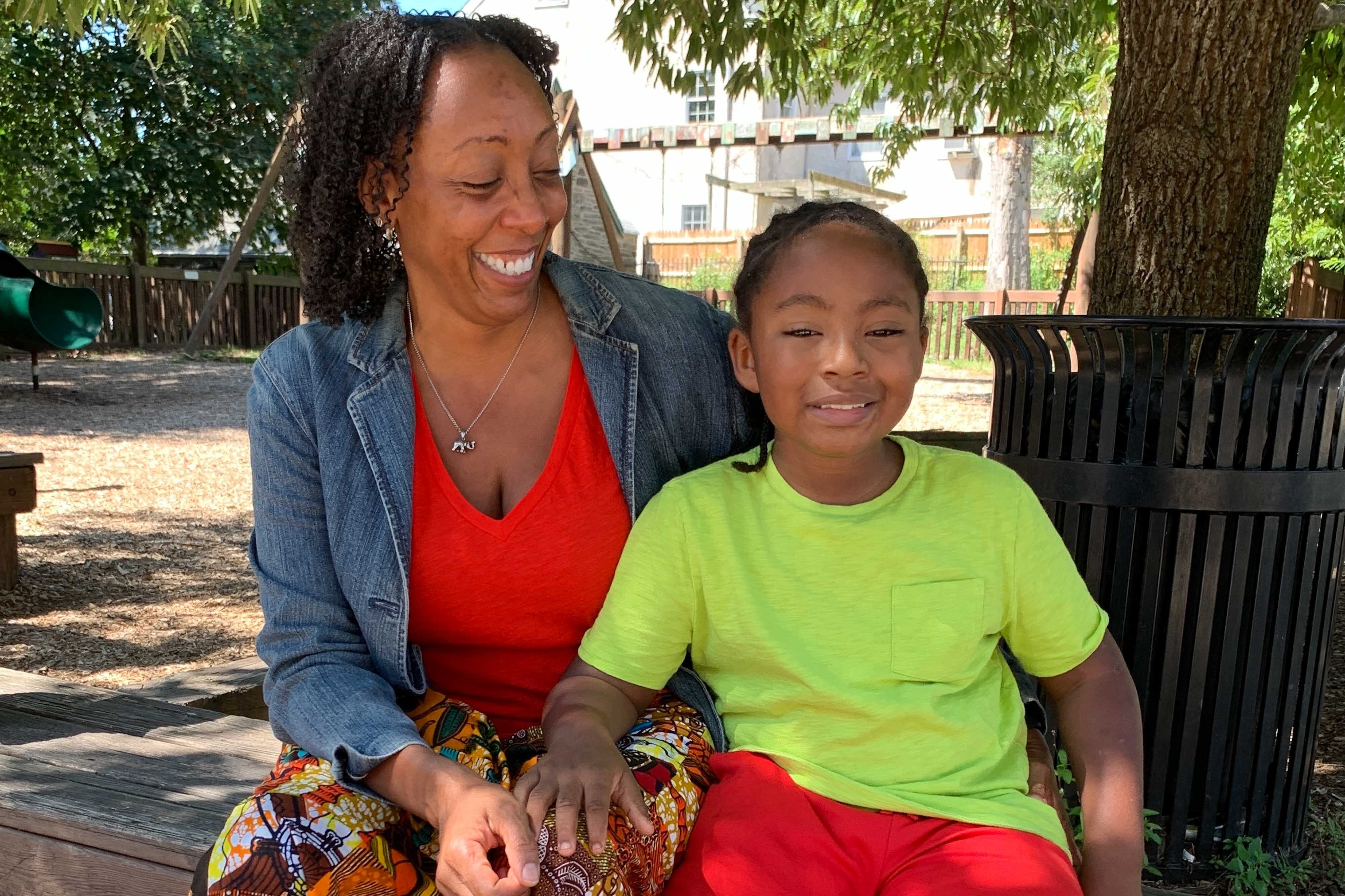 Jeannine Payne sits on a bench with her son Andre. She wears a red top and a denim jacket. He’s wearing a neon green shirt and red shorts.