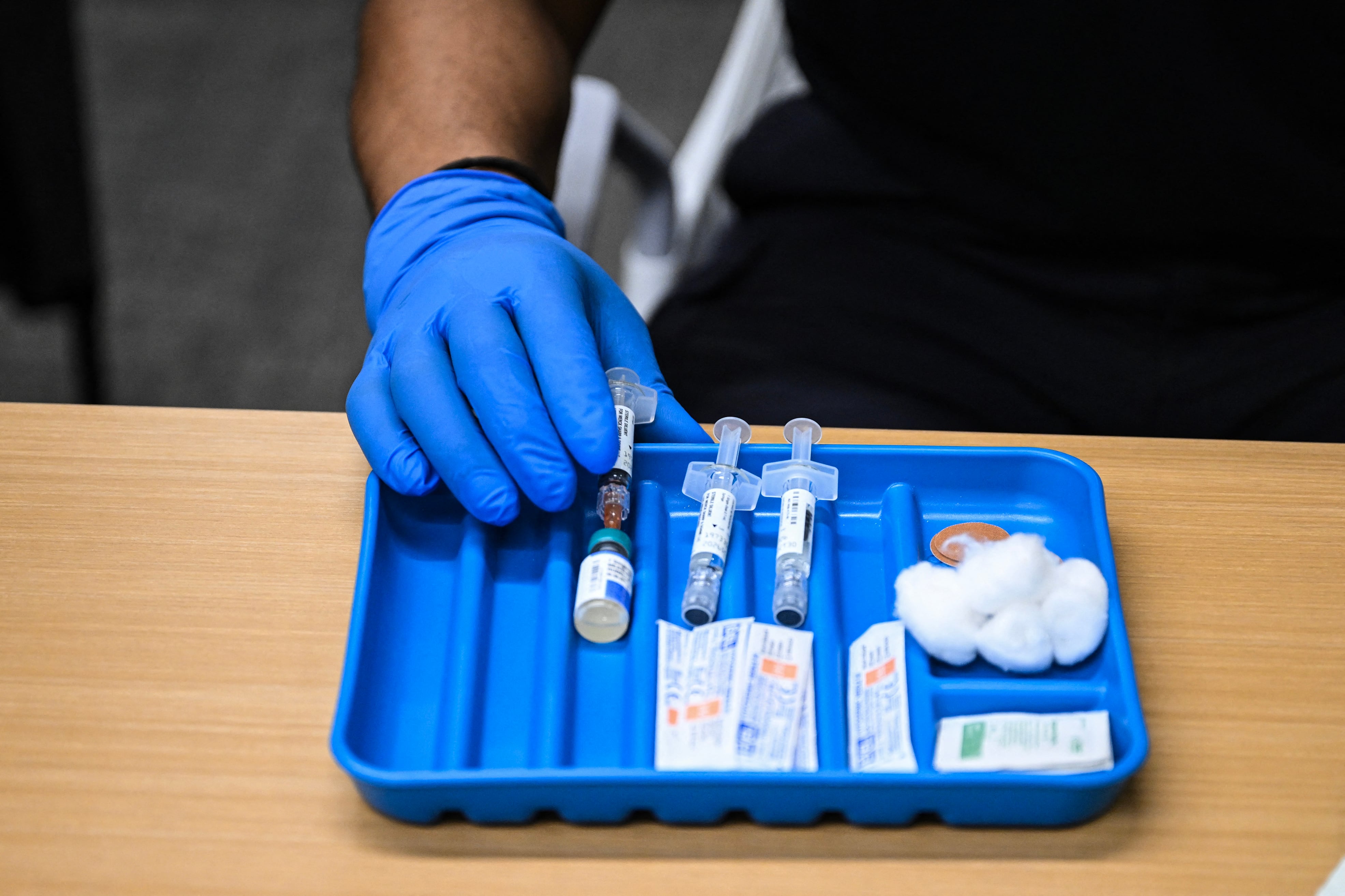 A close up of a single hand with a blue medical glove holding a vile of a vaccine next to cotton balls, syringes and alcohol wipes.