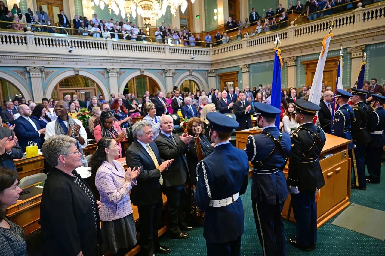 A uniformed color guard presents colors in a packed Colorado House of Representatives. The room is ornately decorated with green walls.