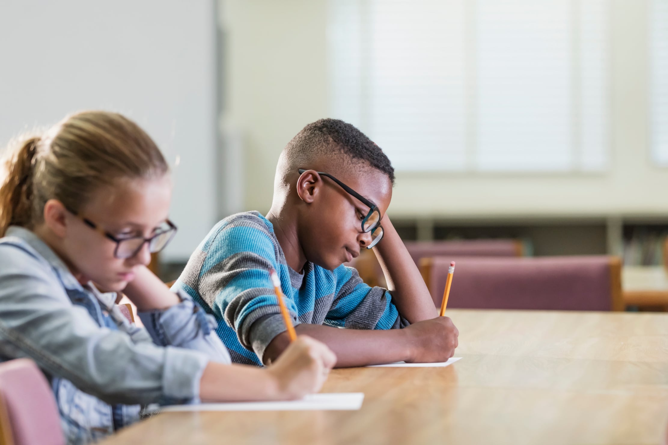 Two young students sit inside of a classroom at a wood table. Both of them are taking an exam with yellow number two pencils.