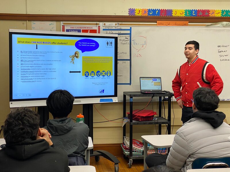 A student wearing a red jacket stands at the front of a classroom with students sitting at desks in the foreground and a whiteboard in the background.