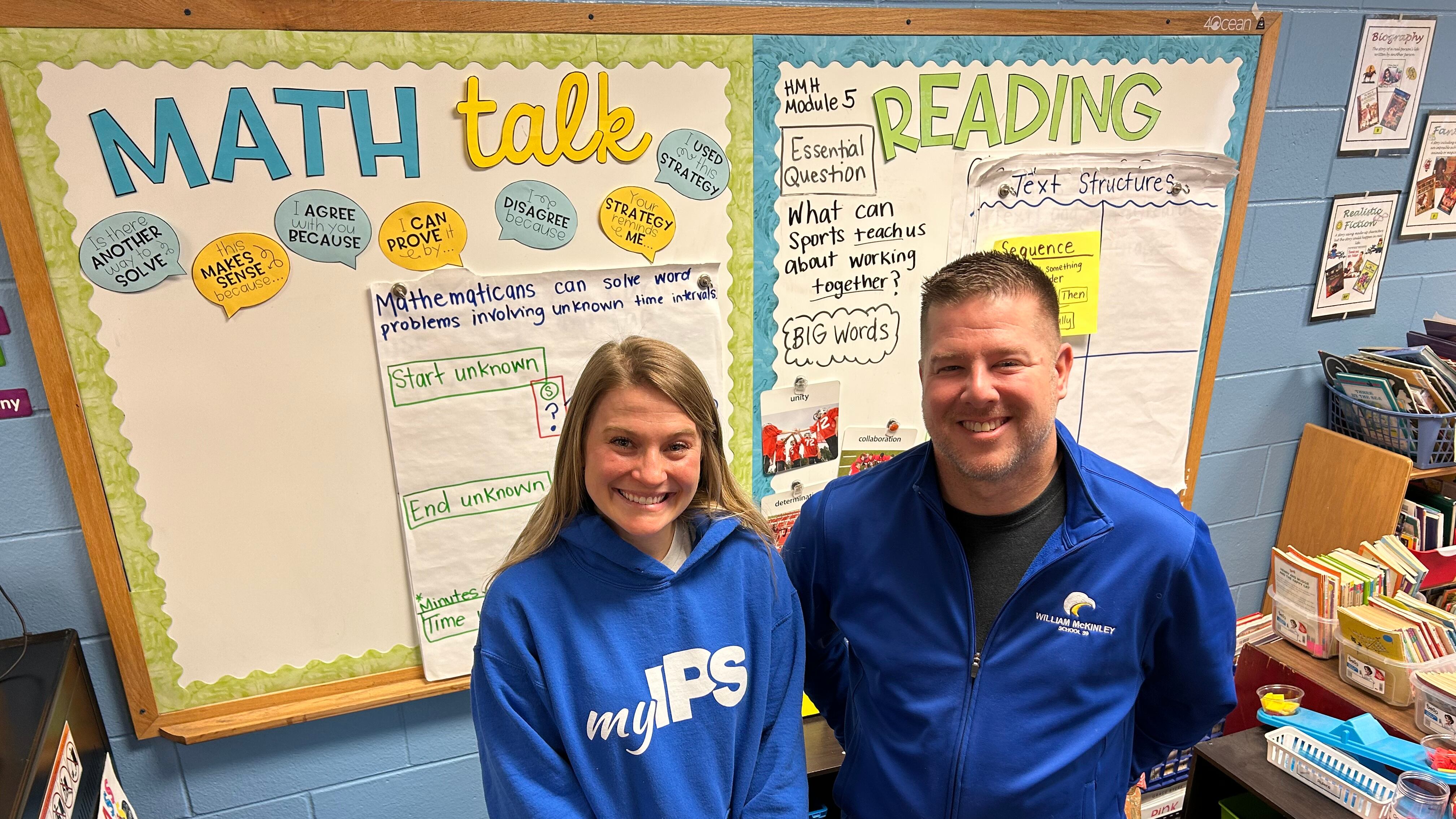 A blonde woman and a man stand smiling in blue shirts in front of two classroom boards, one which says “math” and one which says “reading.”
