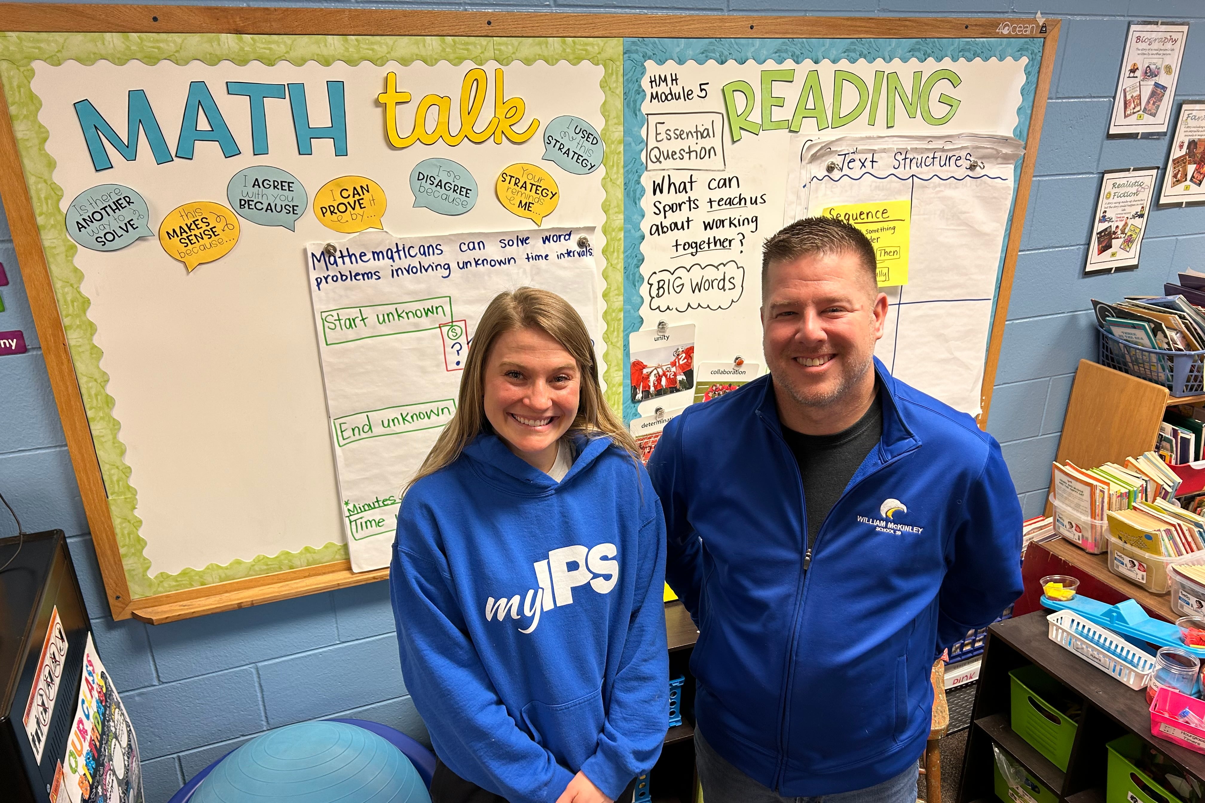 A blonde woman and a man stand smiling in blue shirts in front of two classroom boards, one which says “math” and one which says “reading.”