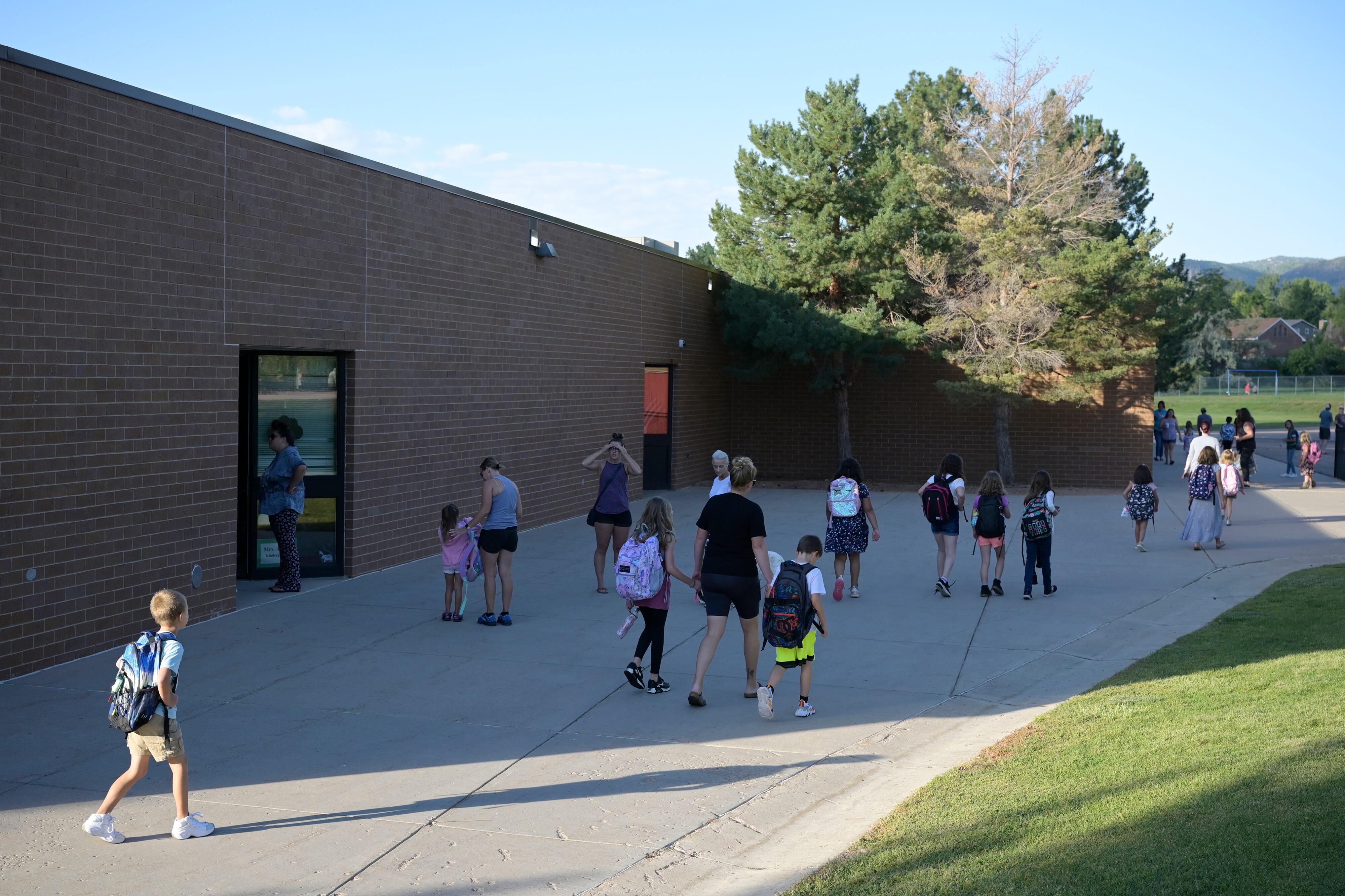 A group of young students and a few adults walk away from the camera next to a brick school building and a blue sky in the background.