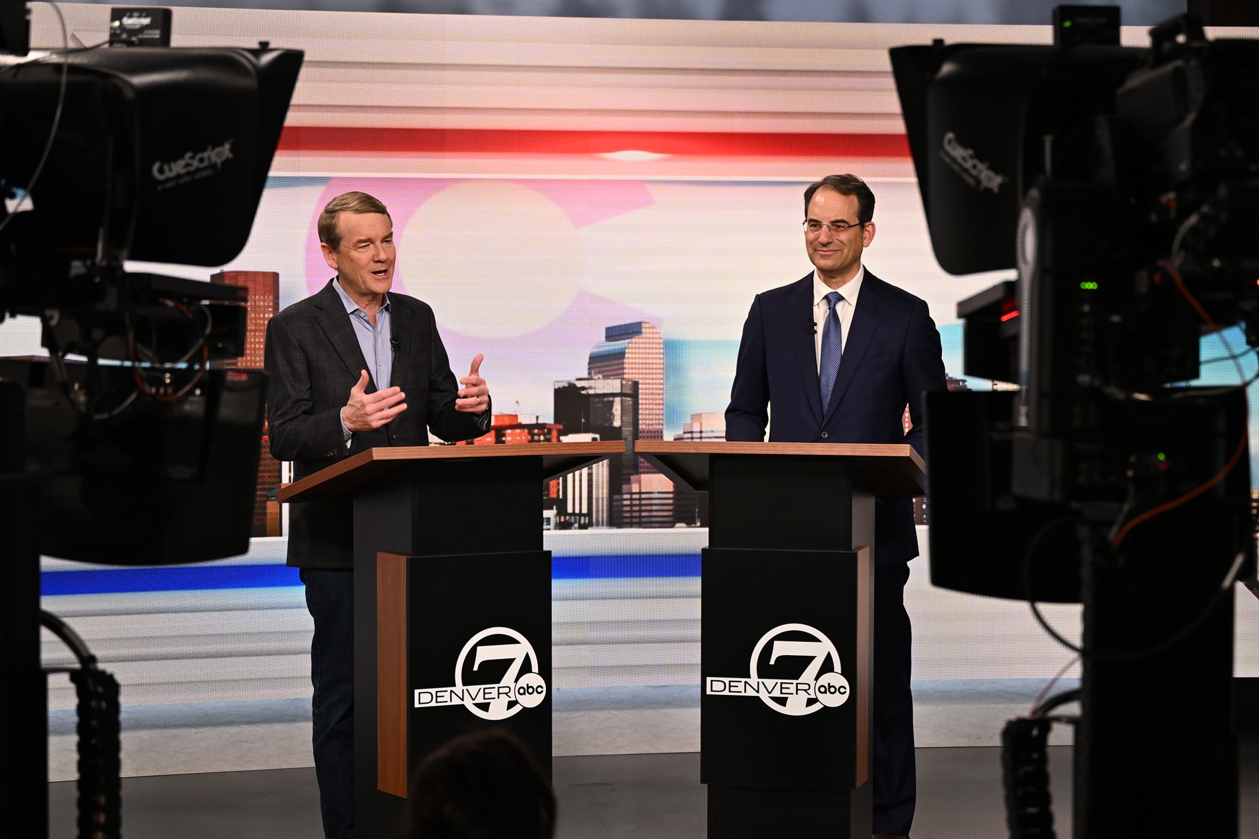 A photograph of two white men in suits speaking from behind podiums on a news cast stage with two cameras on both sides of the photo.