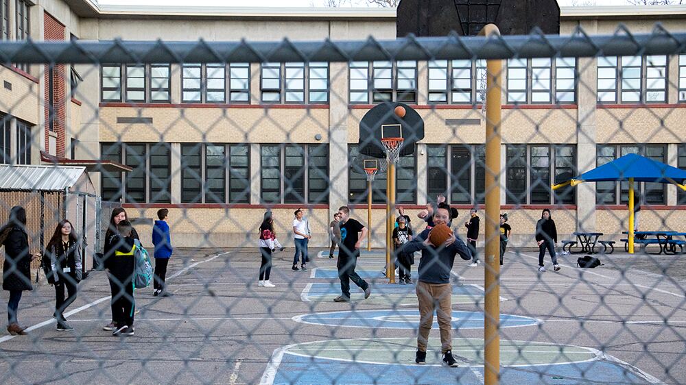Middle school students stand on the blacktop outside a brick school building. They are seen through a chain link fence.