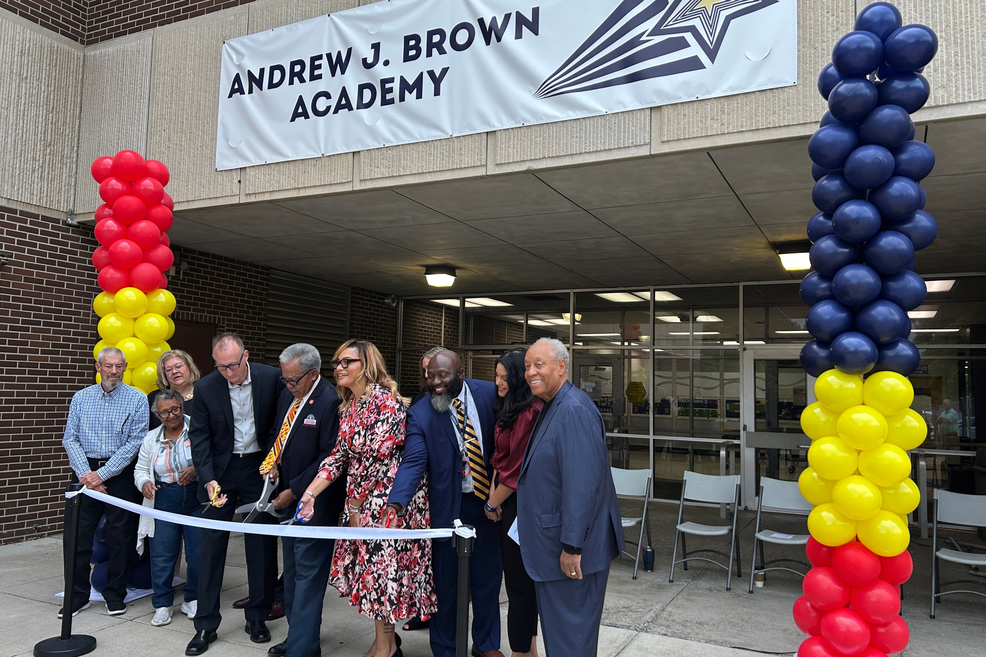 A photograph of a group of adults in business clothes lined up ready to cut a ribbon outside of a school building entrance with balloons on each side.