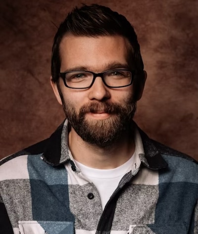 A man with a brown beard and glasses and brown hair wearing a plaid shirt and a white Tshirt. This is a headshot