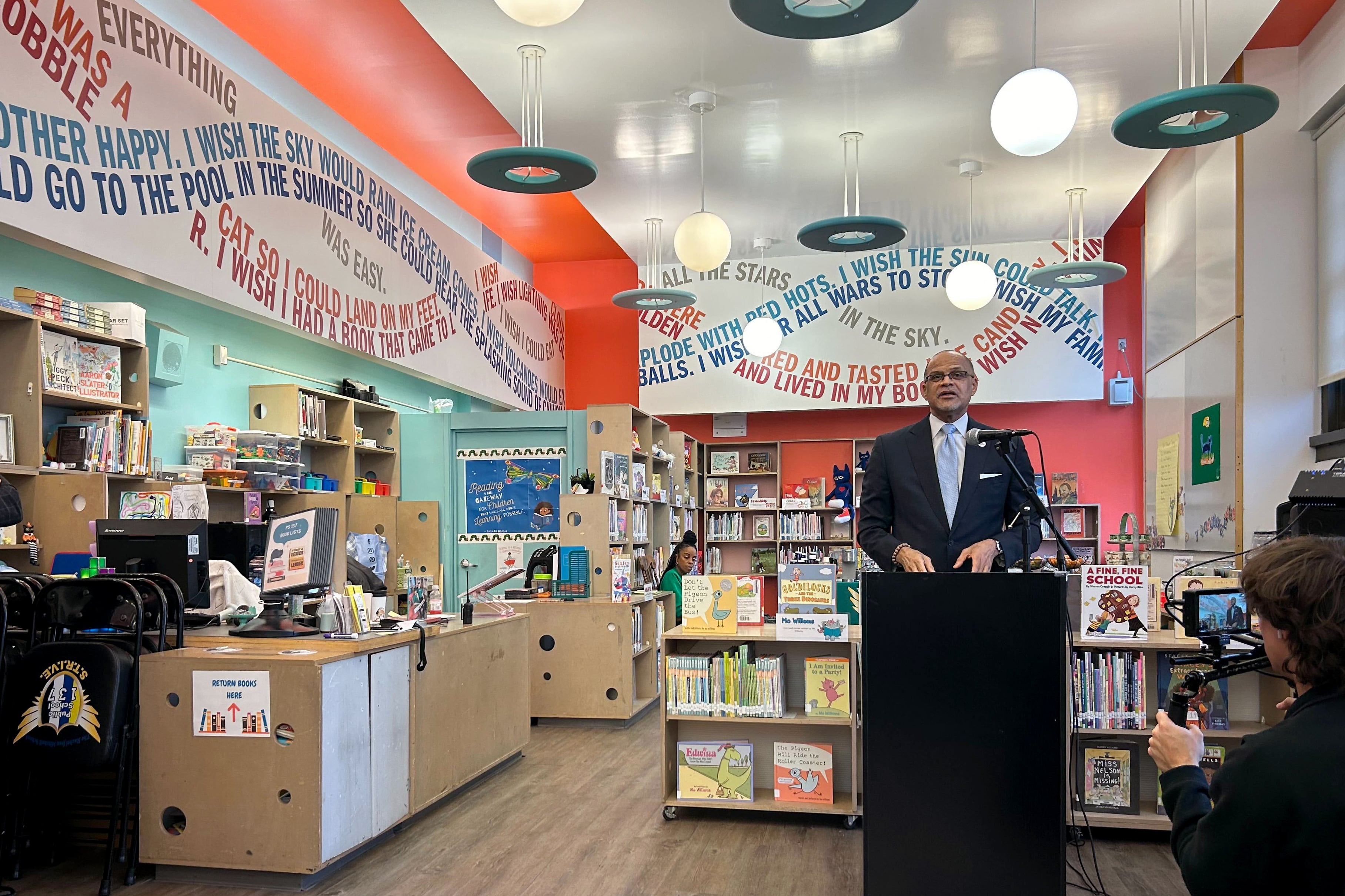 A man wearing a dark suit and glasses speaks from a podium with a library in the background.