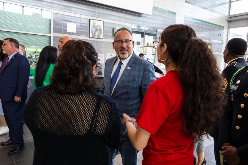 A man wearing a blue suit speaks to two high school students in a school lobby.