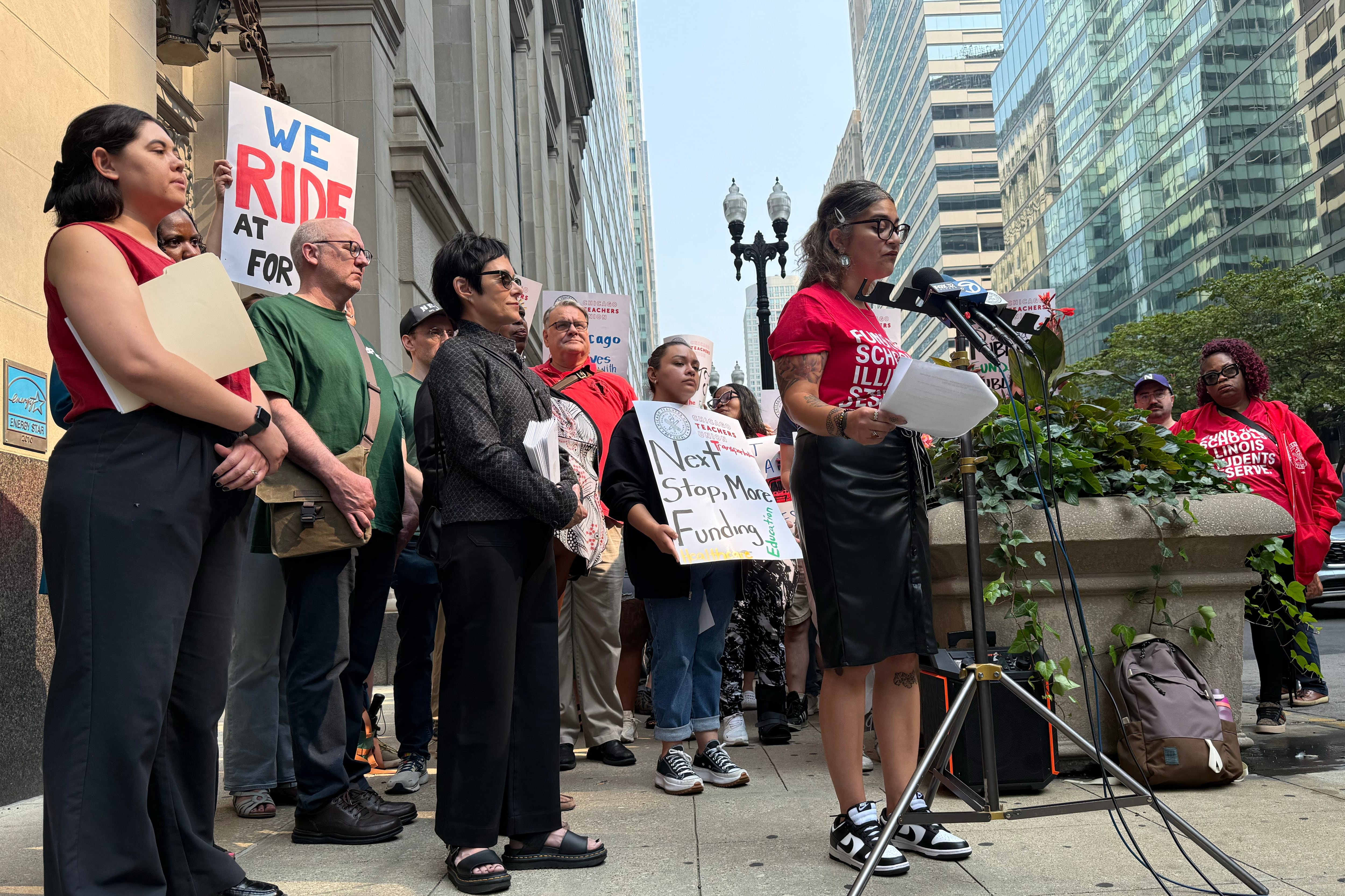 A photograph of a large group of people standing outside, some holding signs, while one person speaks from a microphone.