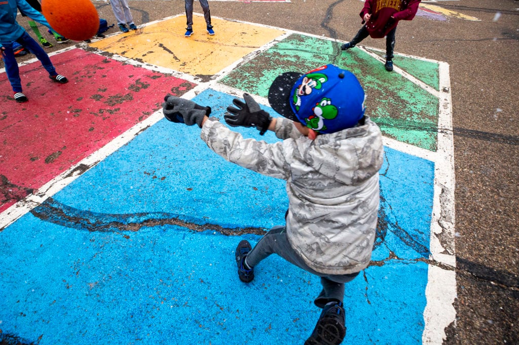 An elementary school child in a sweatshirt, baseball hat, and gloves stands on a foursquare court painted on blacktop on a playground. The child’s back is to the camera and their arms are outstretched as if hitting the ball.