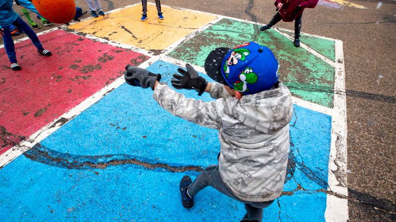 An elementary school child in a sweatshirt, baseball hat, and gloves stands on a foursquare court painted on blacktop on a playground. The child’s back is to the camera and their arms are outstretched as if hitting the ball.