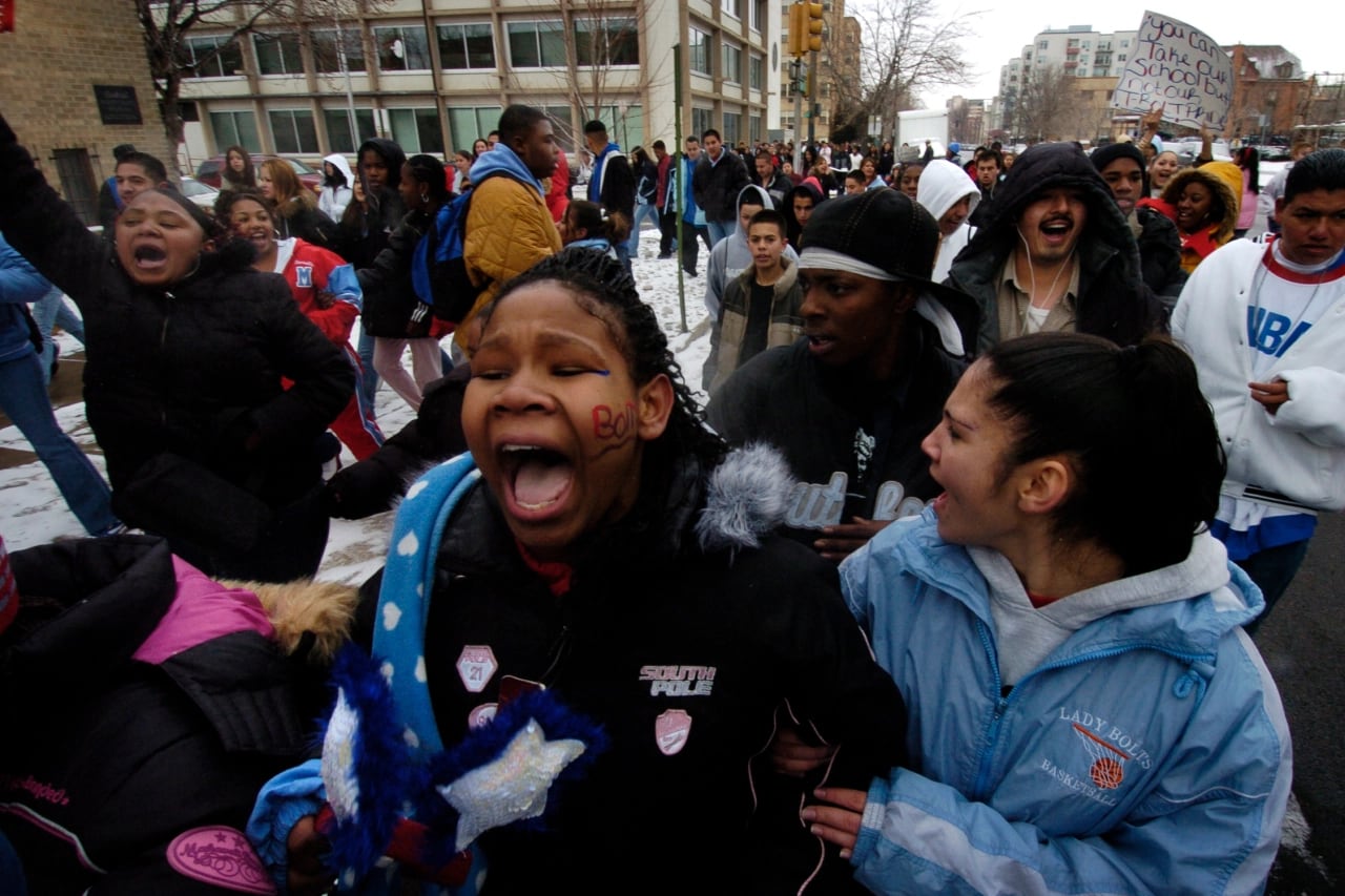 Students shout in protest against the closure of their school. The photo shows a group of students outside in winter. In the center, a girl shouts.