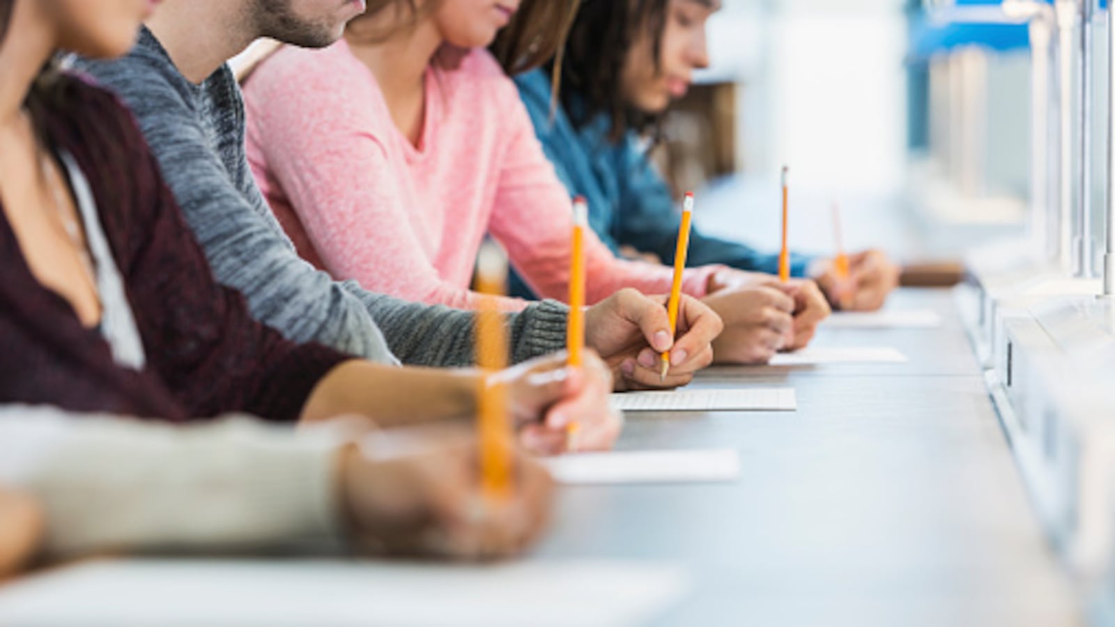 Several students sit at a gray table to take an exam using bright, yellow pencils. The classroom background is white.