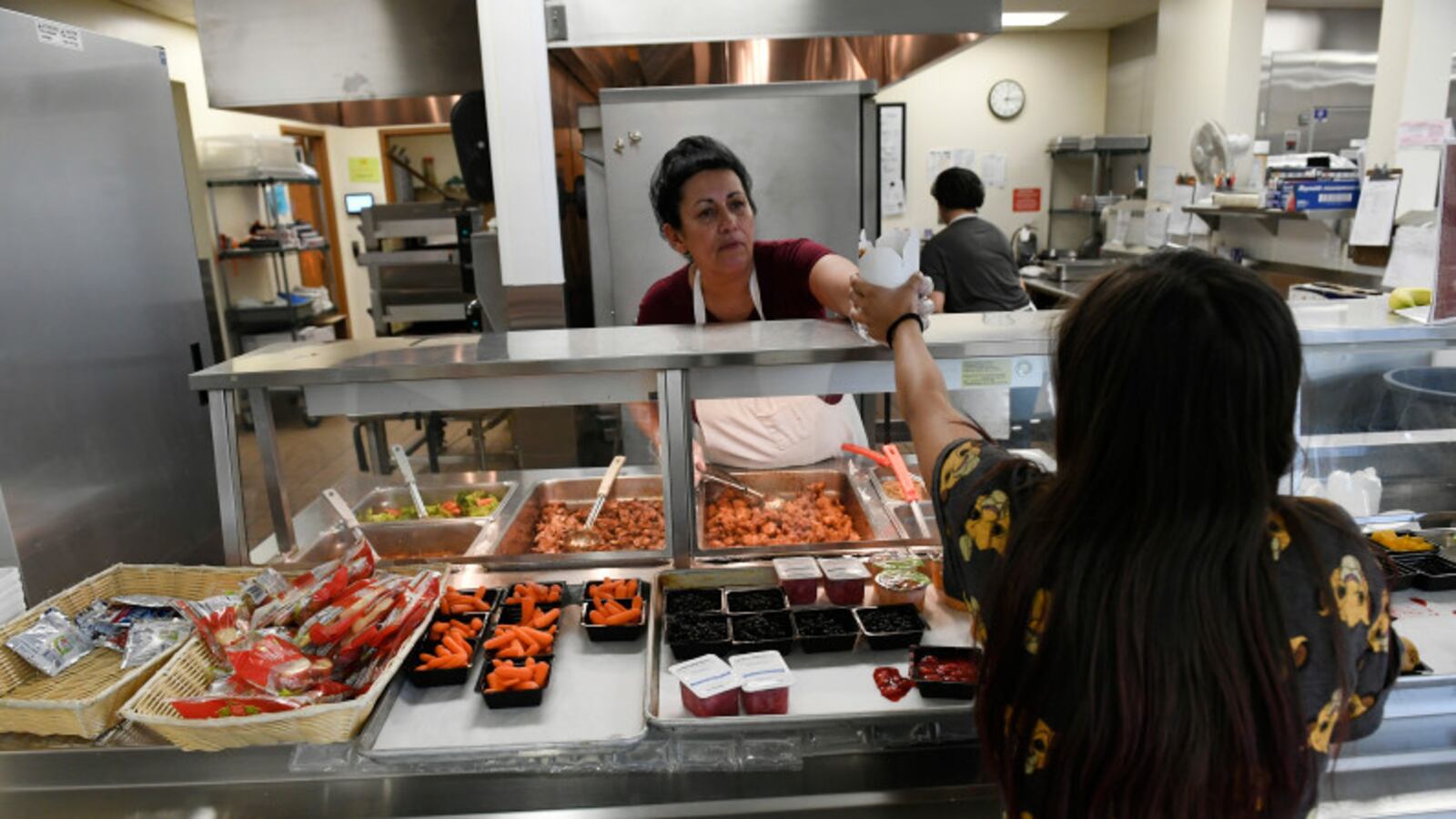 A woman food worker wearing a white apron hands hands a high schooler with long hair a take-out carton from a lunch line, with a kitchen in the background. The worker serves from pans of lunch items, while in the foreground trays feature small containers of carrots, cookies and fruit and other items to grab.