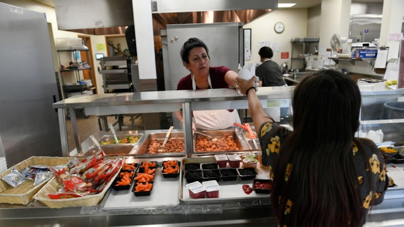 A woman food worker wearing a white apron hands hands a high schooler with long hair a take-out carton from a lunch line, with a kitchen in the background. The worker serves from pans of lunch items, while in the foreground trays feature small containers of carrots, cookies and fruit and other items to grab.