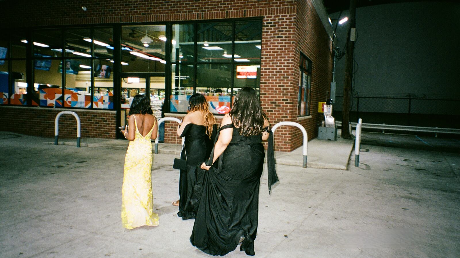 Three young women in prom dresses make their way toward a convenience store.