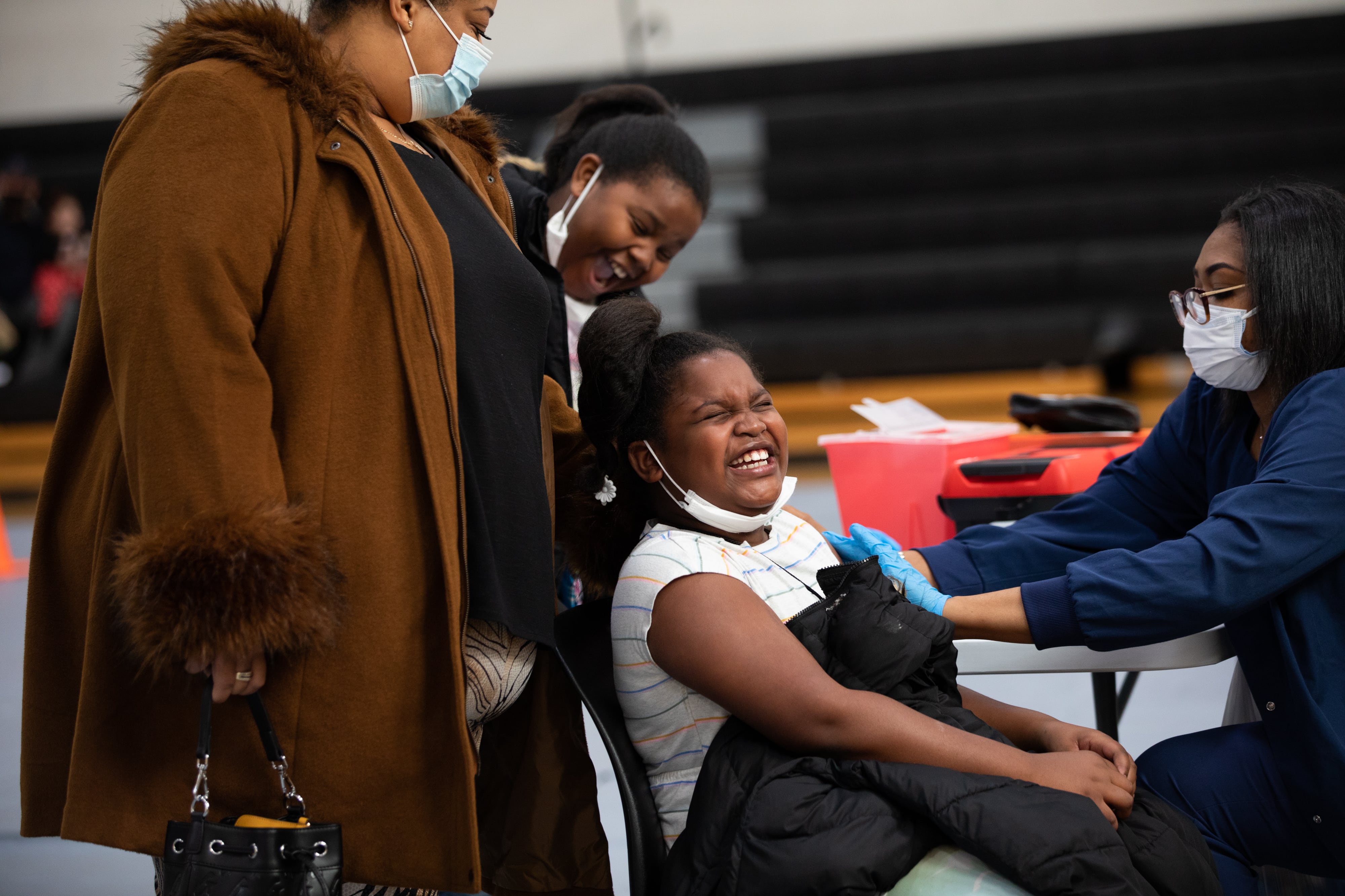 A child laughs as she gets a COVID-19 vaccine while another child and an adult hover above her.