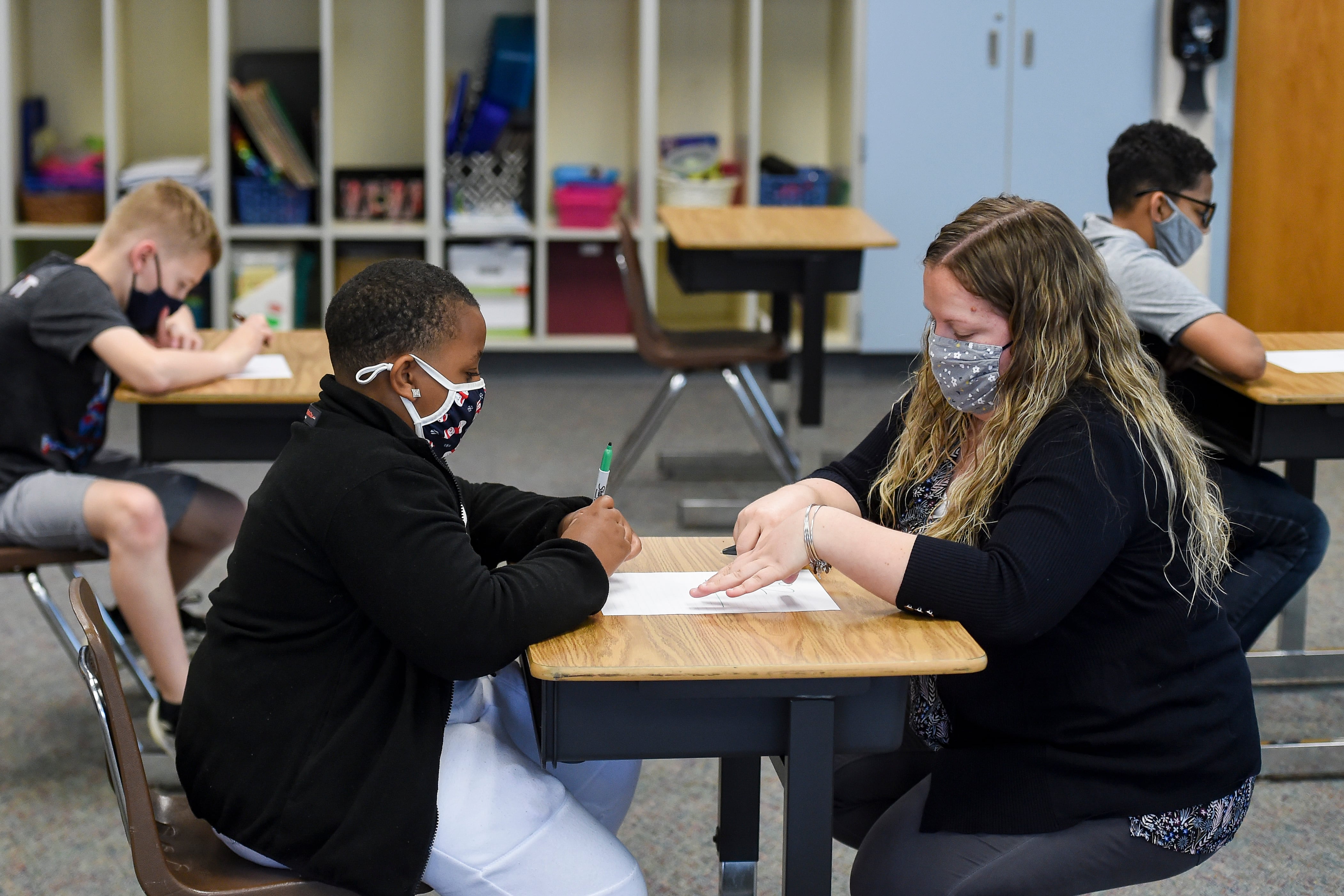 A student wearing a face mask and his teacher sit across from each other at his desk in a classroom.