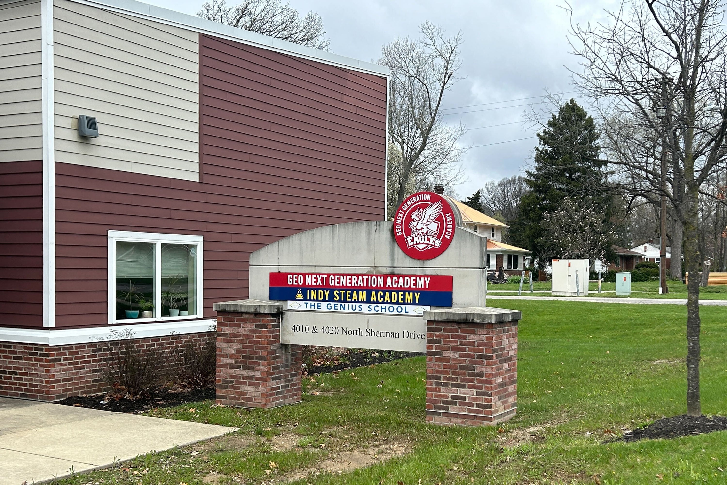A stone sign with red, white, yellow and blue writing sits on a grassy lawn next to a brick building.