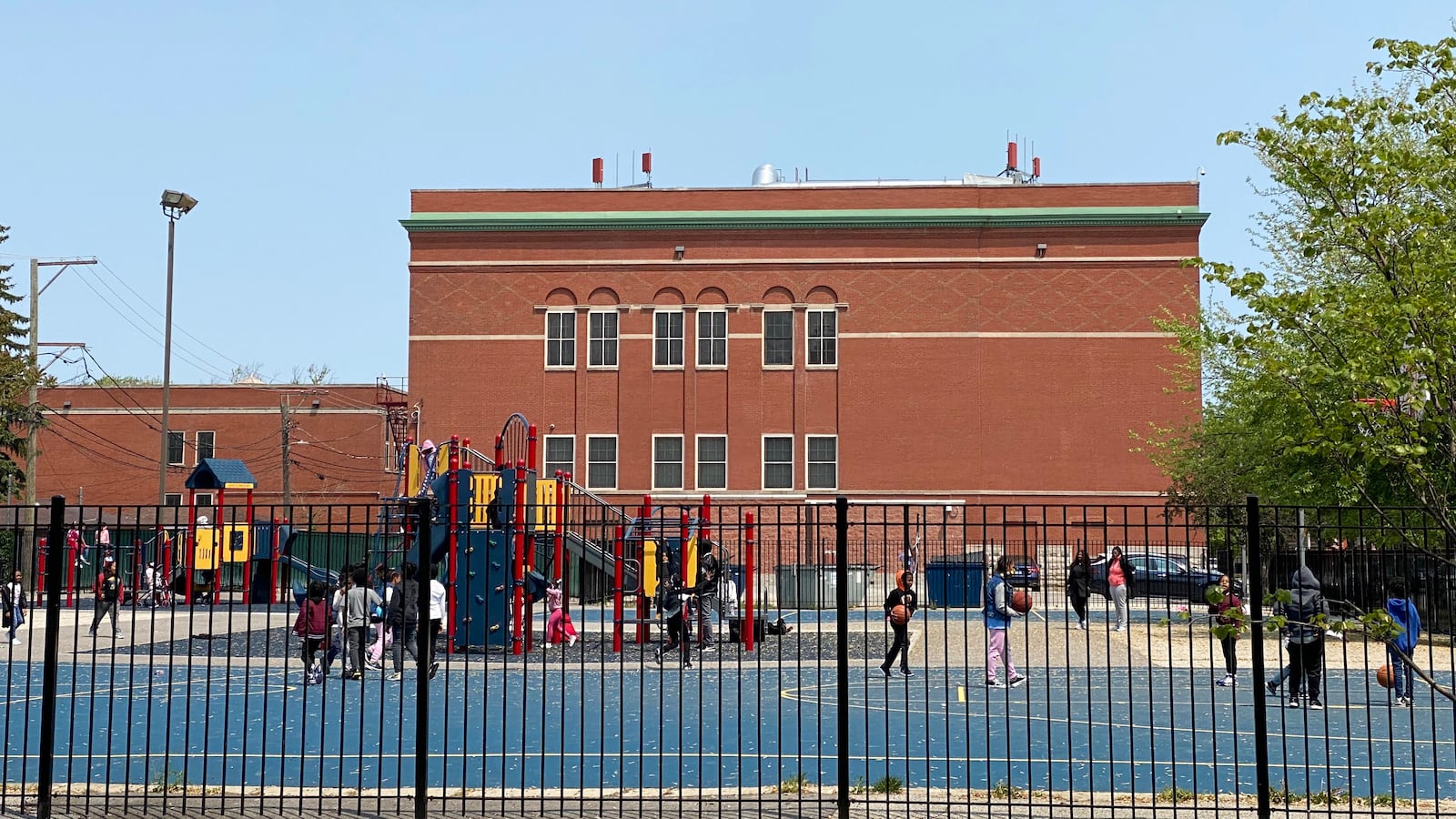 Behind a large fence, a group of young students play on a school playground with a large red brick building in the background.