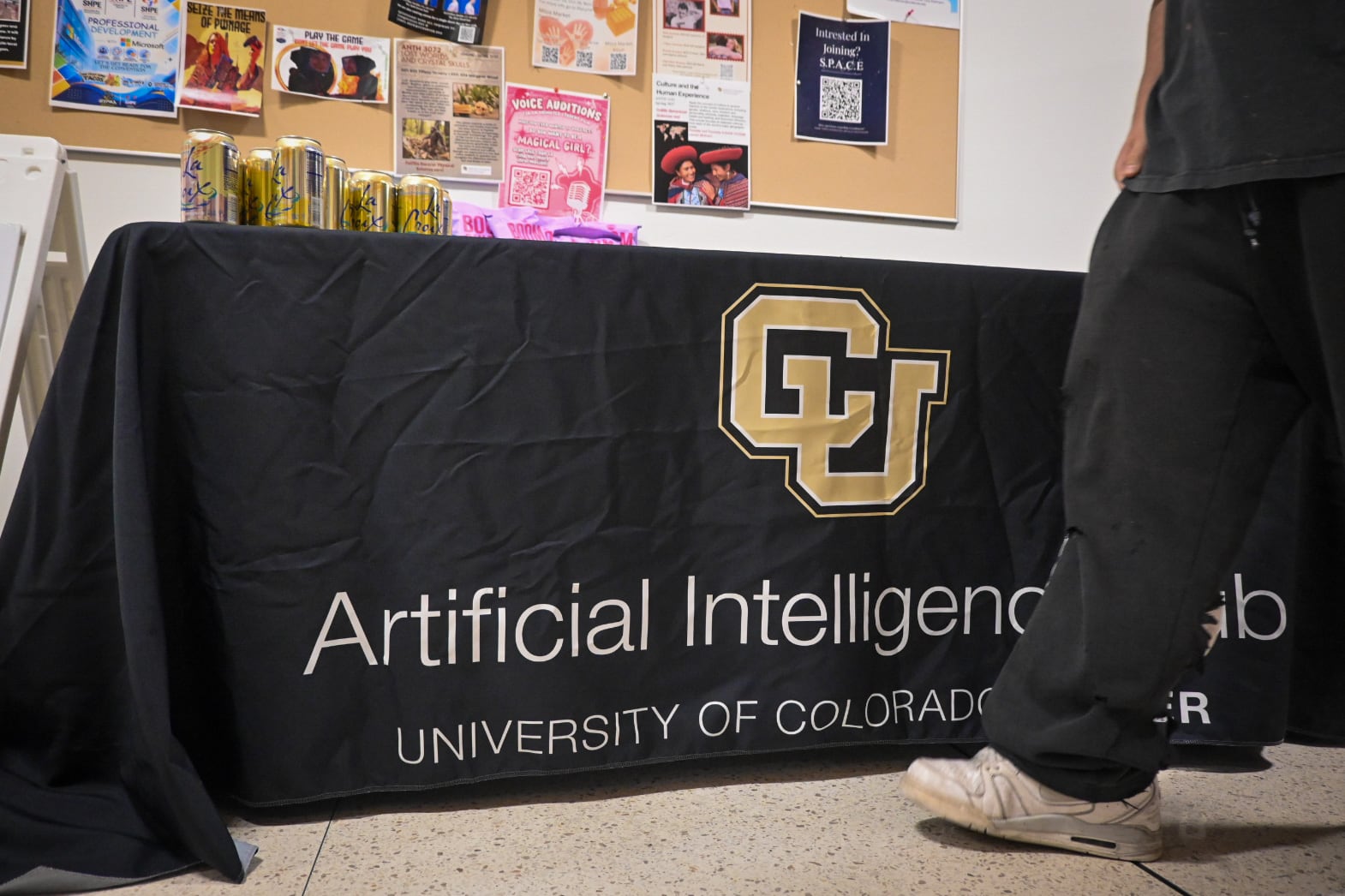 A black tablecloth that says CU Artificial Intelligence Hub is draped over a table as a person in a black shirt, black pants, and white shoes walks by.