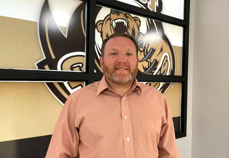 A man in a peach shirt stands in front of school mascot.