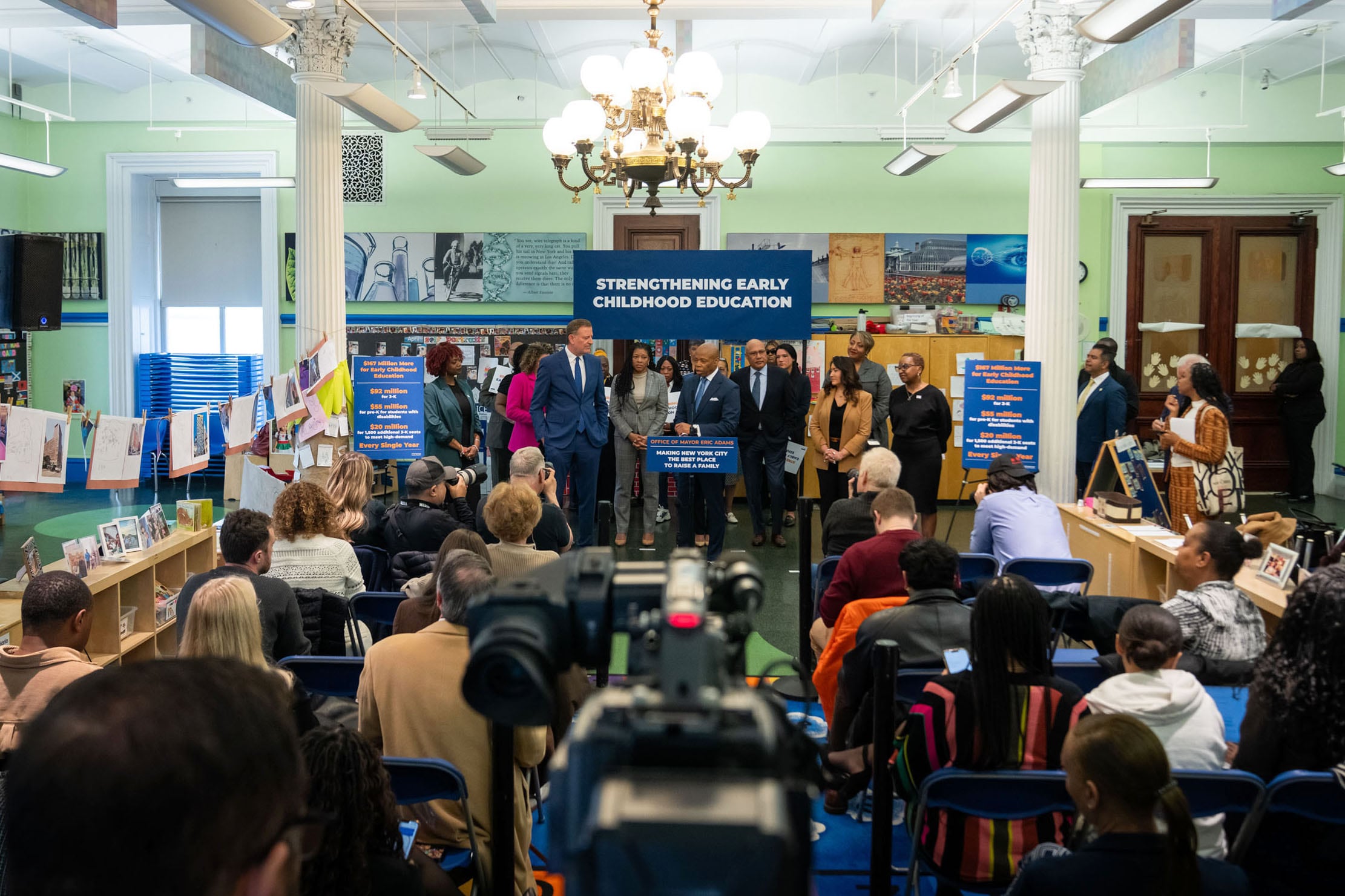 A group of adults in suits and business clothes stand behind a podium and in front of a large crowd of adults sitting in chairs listening to the press conference.