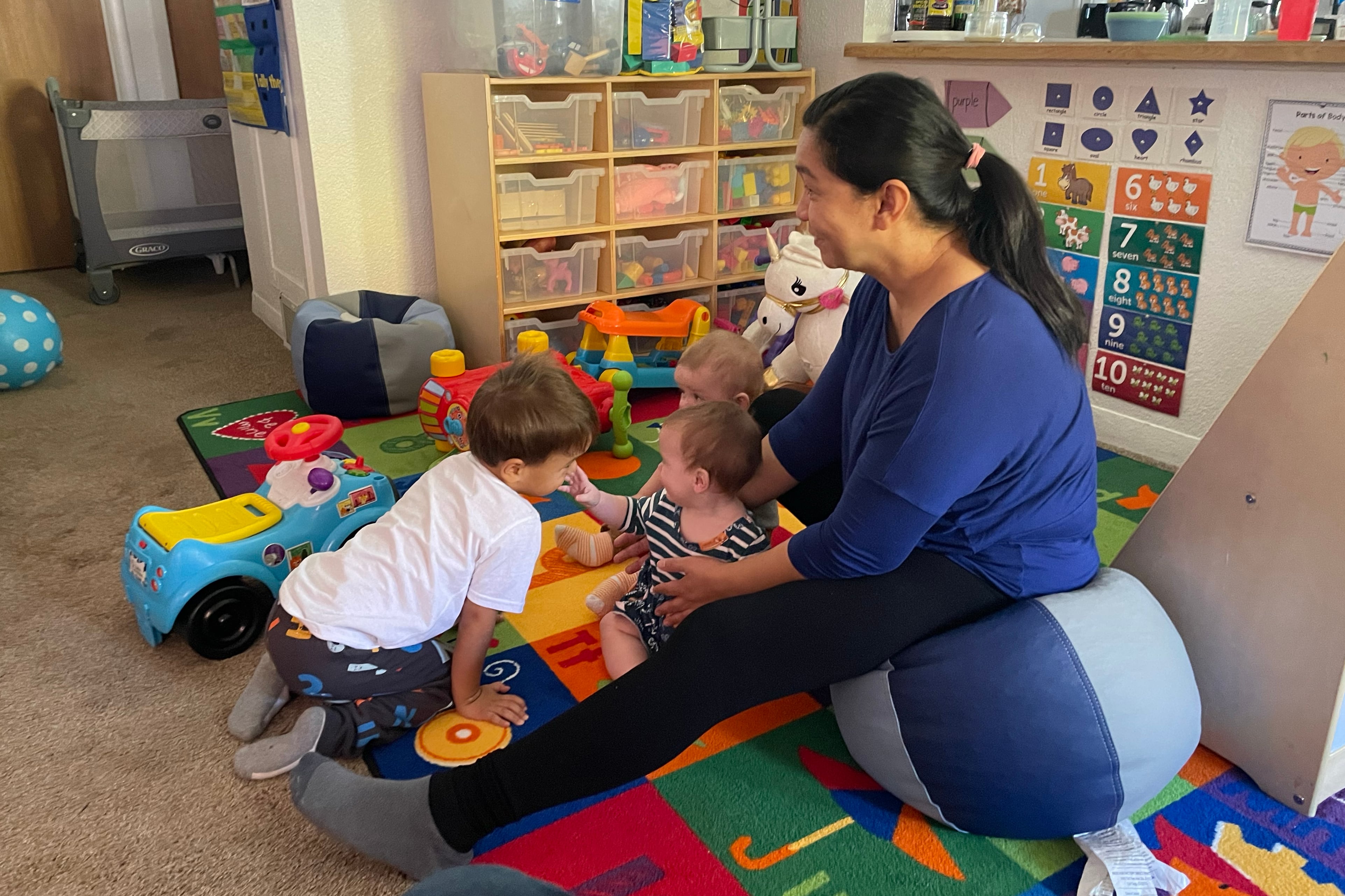 A woman with long dark hair and wearing a blue shirt supports two young children while an older child plays near by.