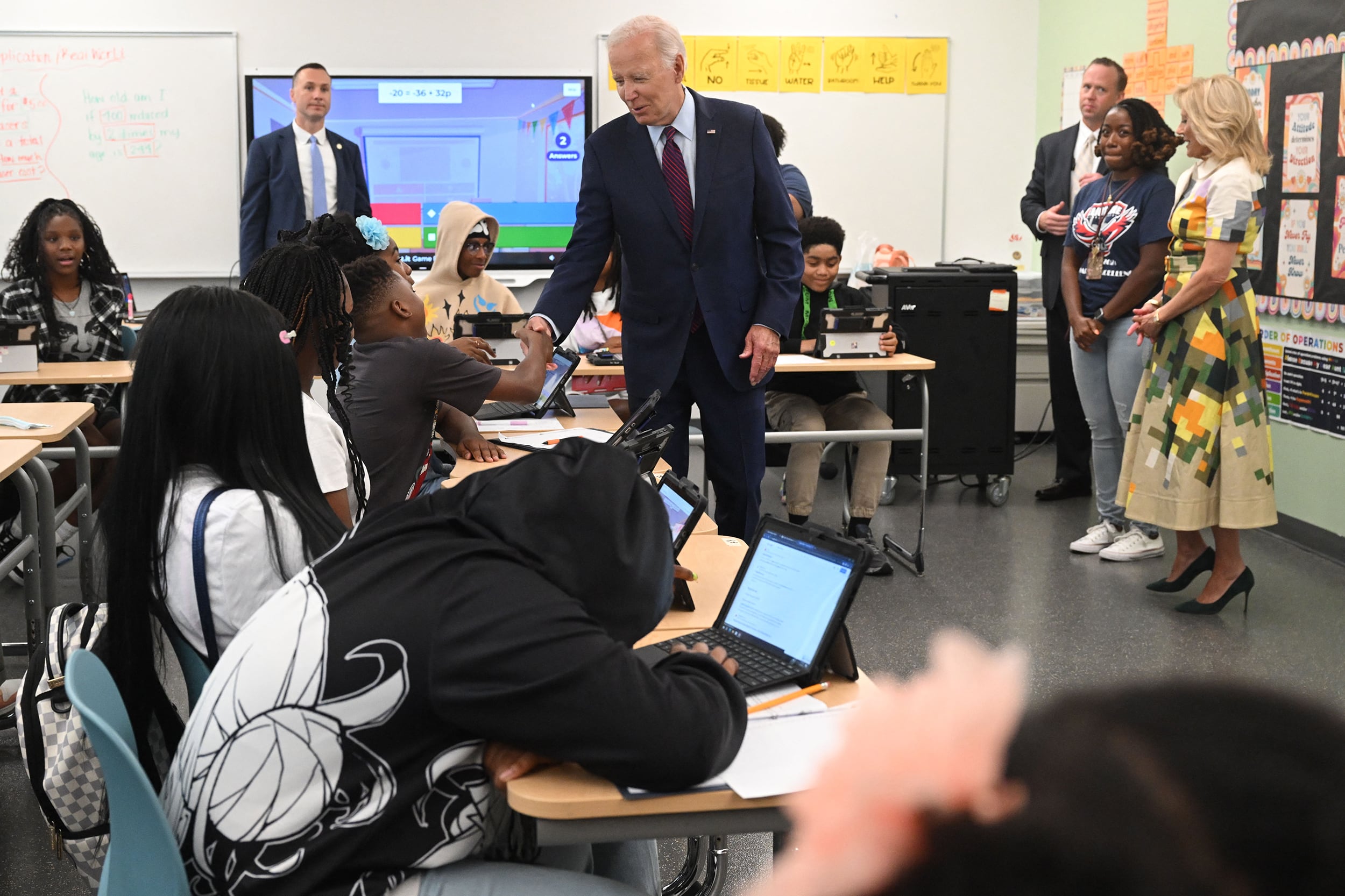 A photograph of a high school classroom with students sitting at desks and five adults standing in the room. One adult, is President Joe Biden in a suit shaking the hand of one student.