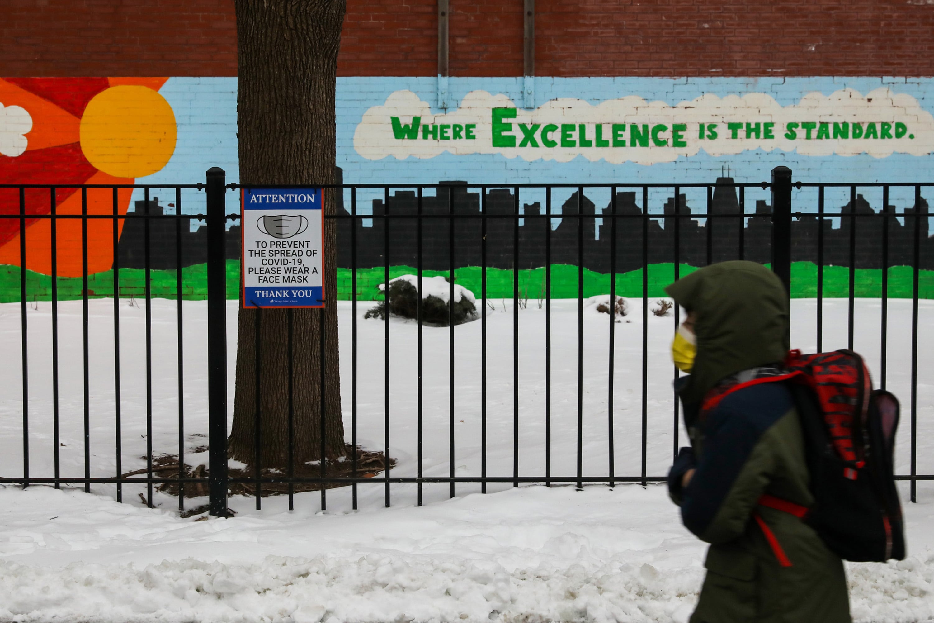 A student, wearing a mask, hooded winter jacket and backpack, walks past a school mural that reads “Where excellence is the standard” and a sign calling for people to wear a mask.