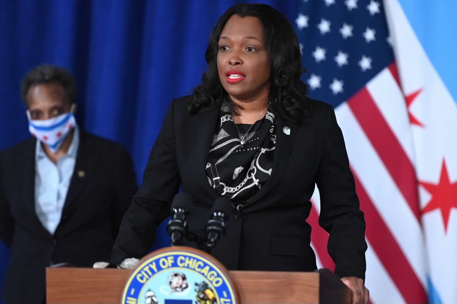 Schools chief Janice Jackson stands at a podium while giving an address announcing her departure. To the left, in the backgrond, is Mayor Lori Lightfoot wearing a mask. To the right are overlapping American and Chicago flags.